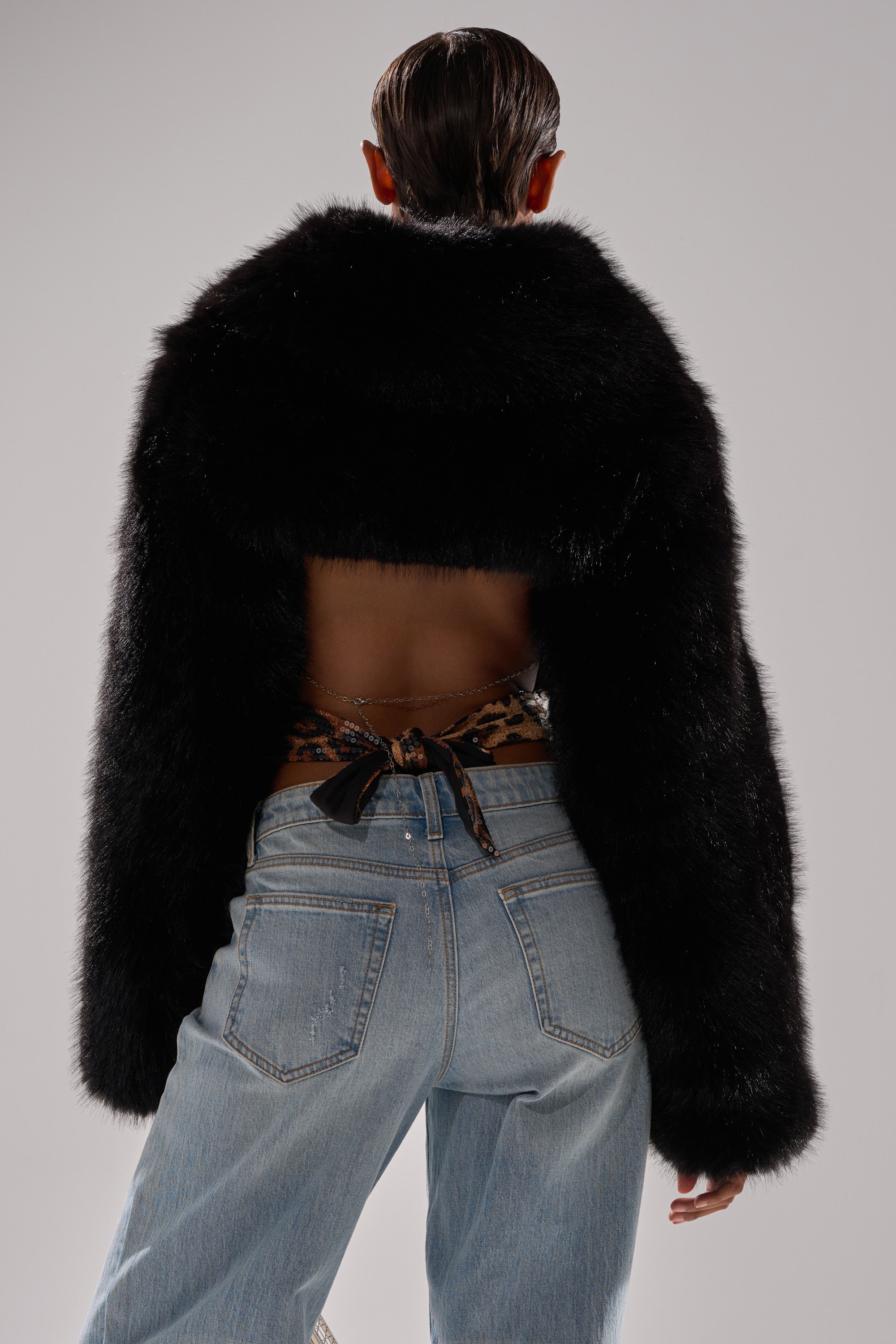 A person faces away, wearing the LEILA ULTRA CROP FAUX FUR IN BLACK jacket that exposes their back, a patterned garment tied at the waist, and light blue jeans against a plain, light backdrop.