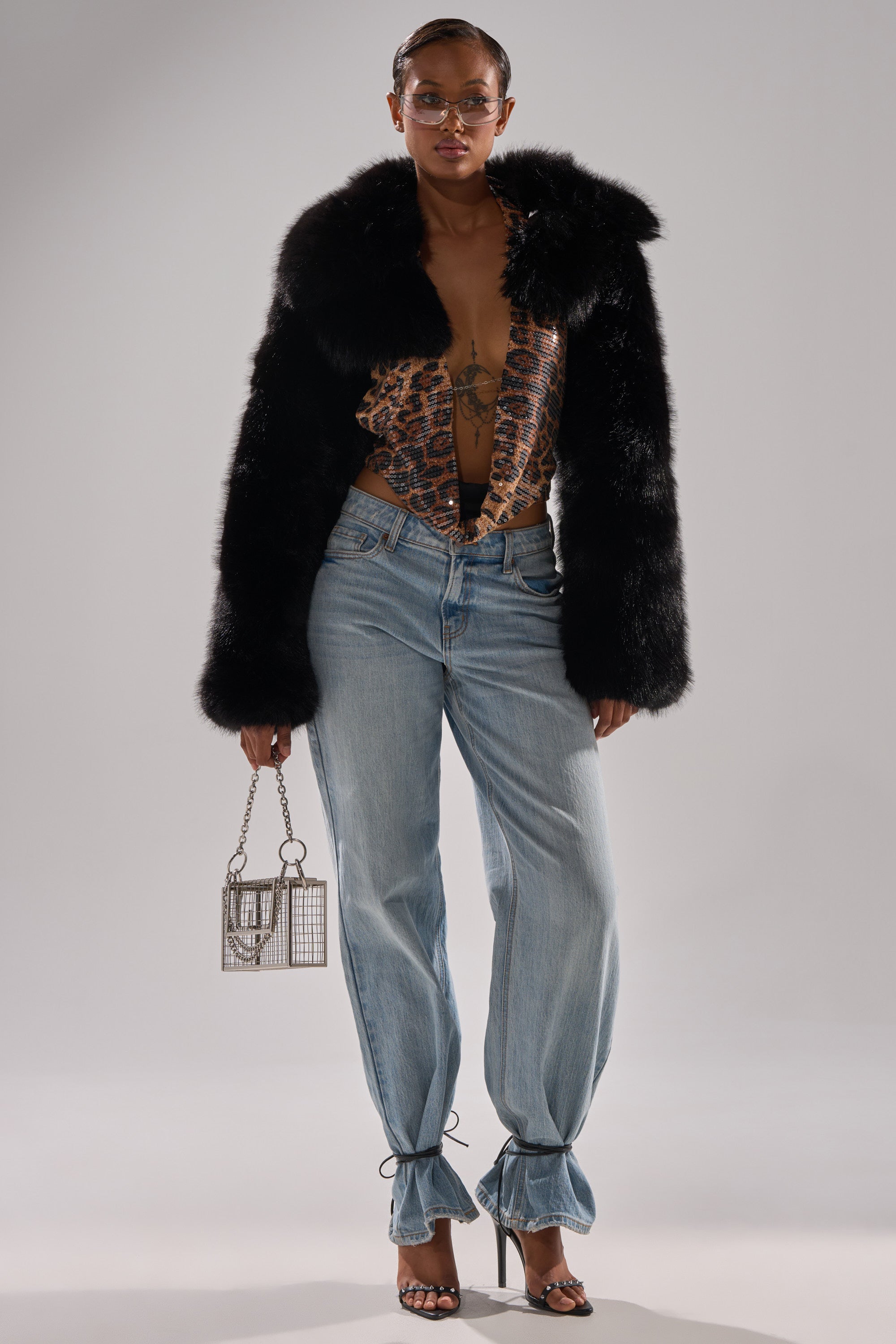 A woman wears the LEILA ULTRA CROP FAUX FUR IN BLACK, paired with a patterned top, light blue ankle-tie jeans, heeled sandals, a small metal cage handbag, and glasses, standing before a plain light background.