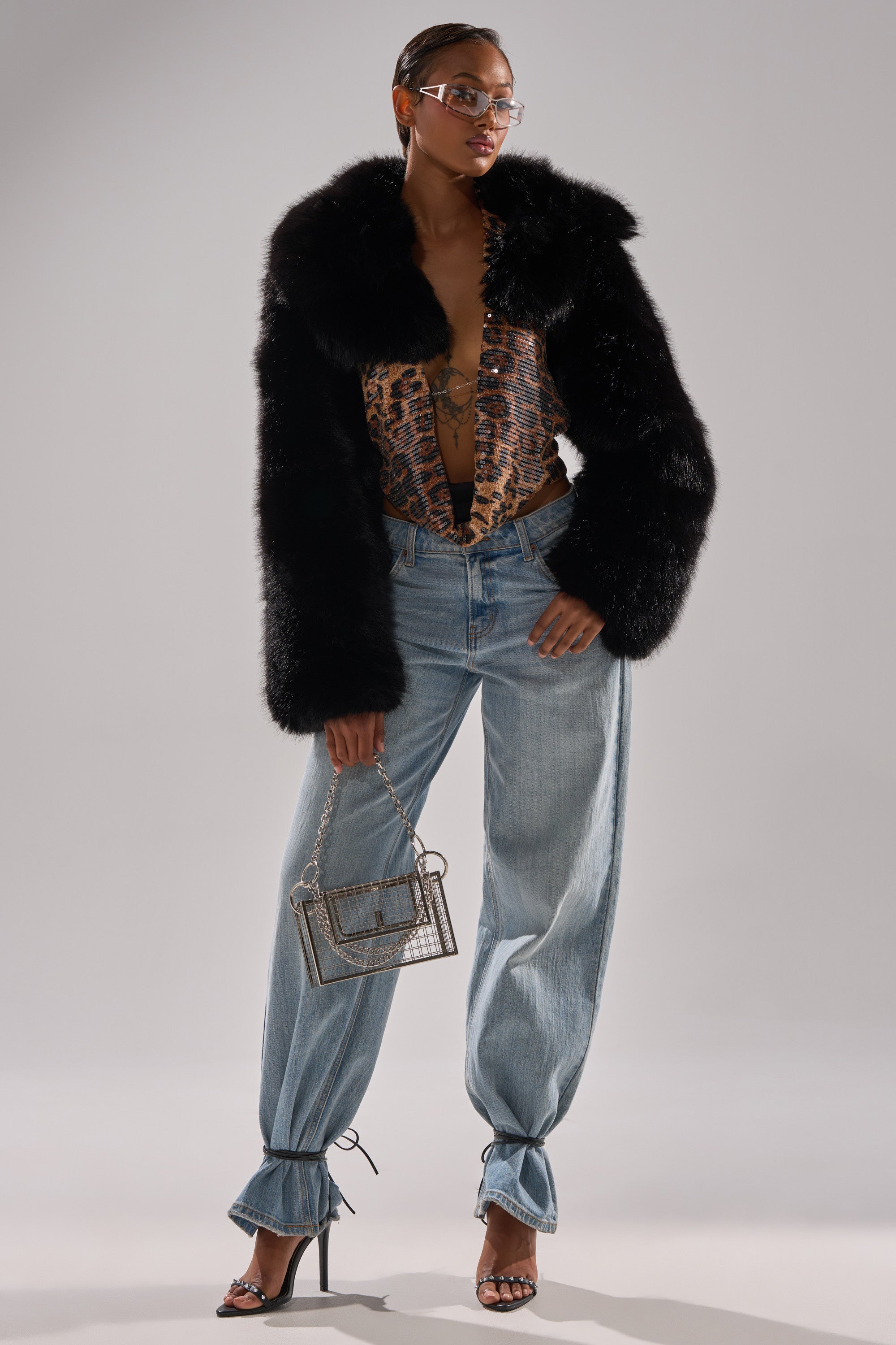 A stylish woman poses in the LEILA ULTRA CROP FAUX FUR IN BLACK, snake print top, ankle-tied blue jeans, strappy heels, clear glasses, and a small transparent handbag.