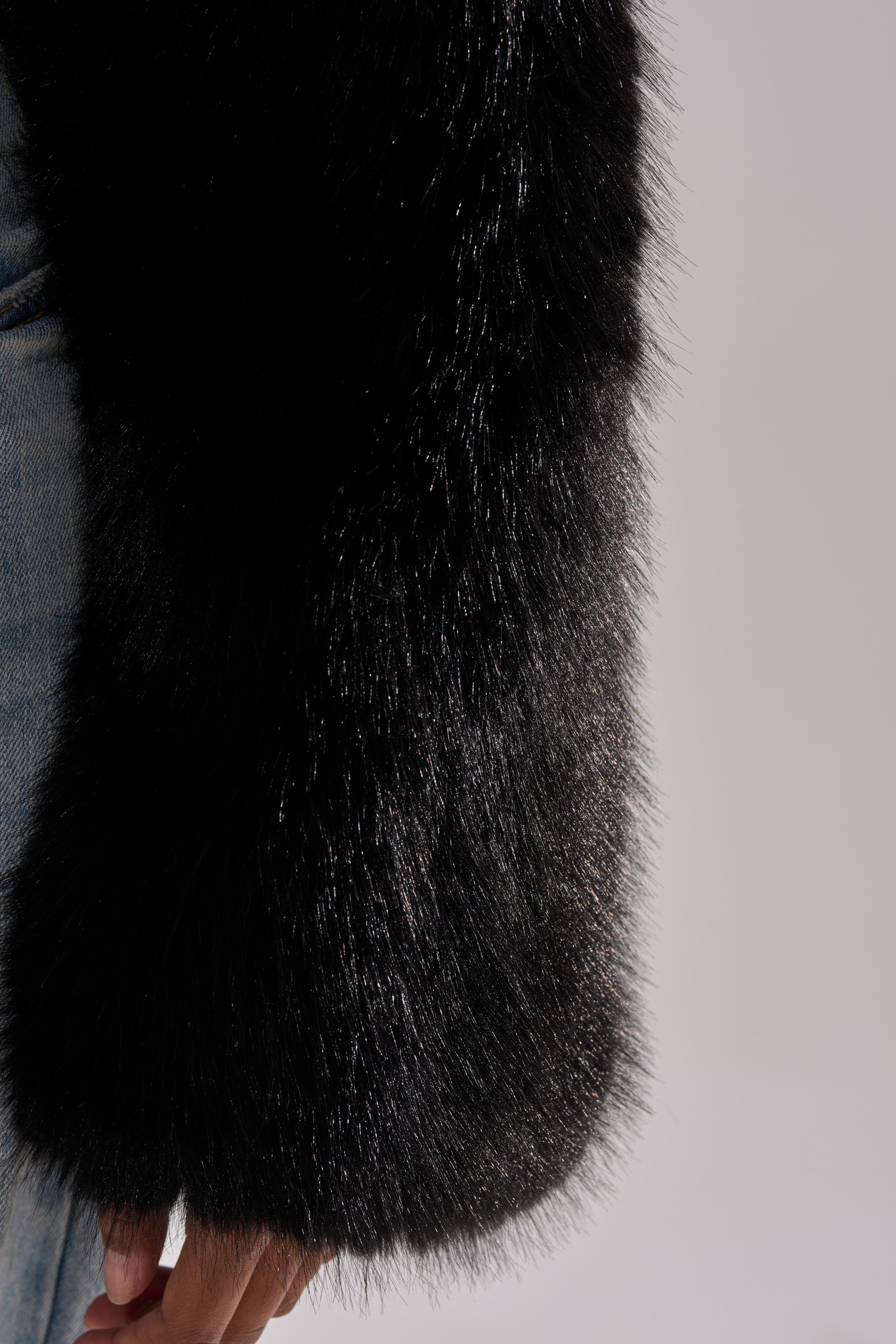 A close-up of a person wearing the LEILA ULTRA CROP FAUX FUR IN BLACK jacket, paired with blue denim jeans, shows their arm and partially visible hand in the frame.