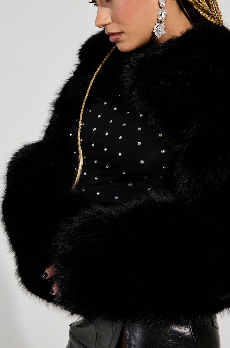 A person dressed in the LEILA ULTRA CROP FAUX FUR IN BLACK jacket over a black, studded top and leather pants. They have long braided hair, a trendy style, and are adorned with large, decorative earrings against a plain white background.