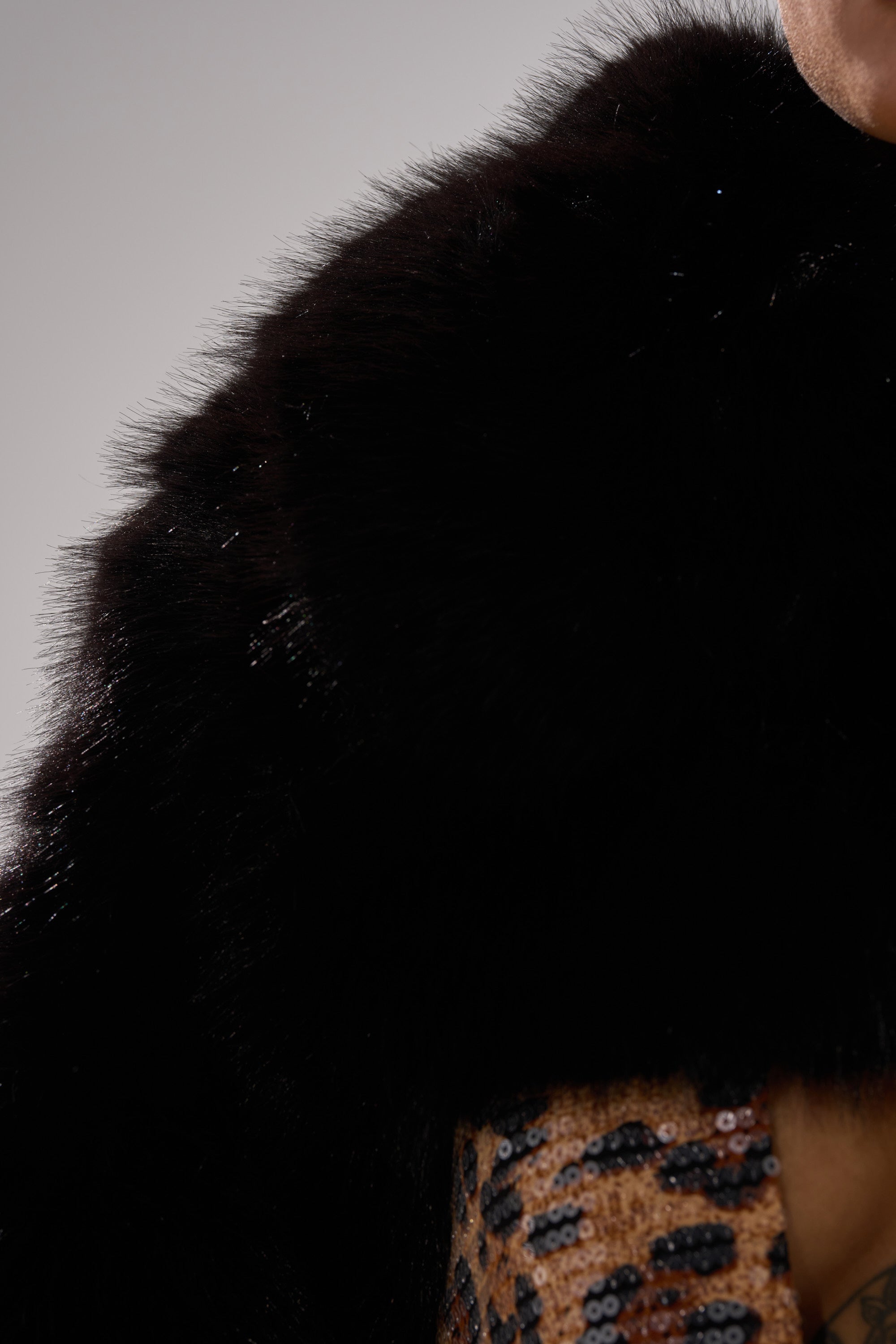 A person wears the LEILA ULTRA CROP FAUX FUR IN BLACK over a sequined animal-print outfit. The neutral background highlights the shawl’s luxurious texture and bold fabric details.