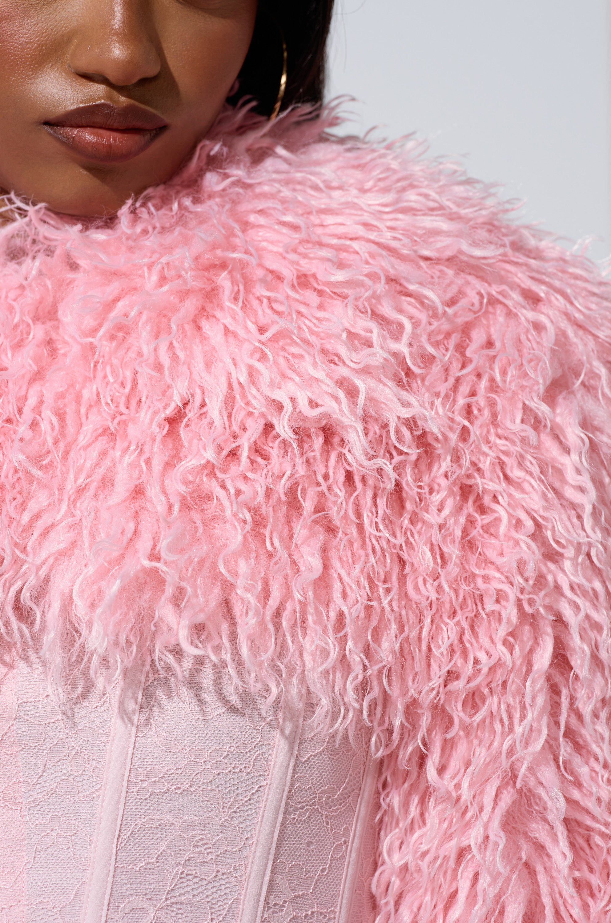 A close-up shows a person wearing the LEILA ULTRA CROP FAUX FUR IN LIGHT PINK over a light pink lace corset, with part of their face and shoulder visible. The jacket features a curly, voluminous faux fur texture.
