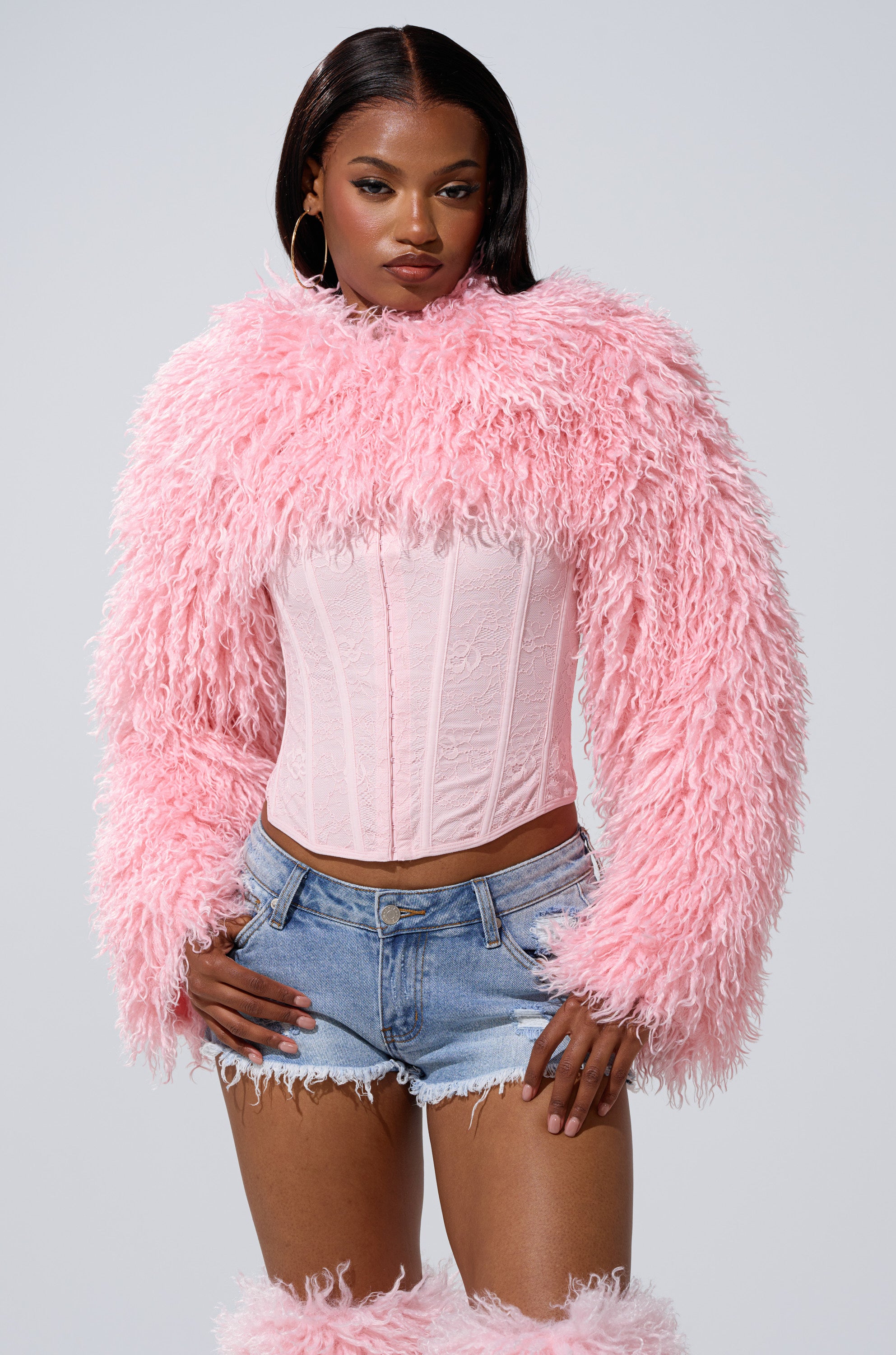 A woman poses with one hand on her hip against a plain background, wearing the LEILA ULTRA CROP FAUX FUR IN LIGHT PINK over a light pink corset and blue denim shorts.