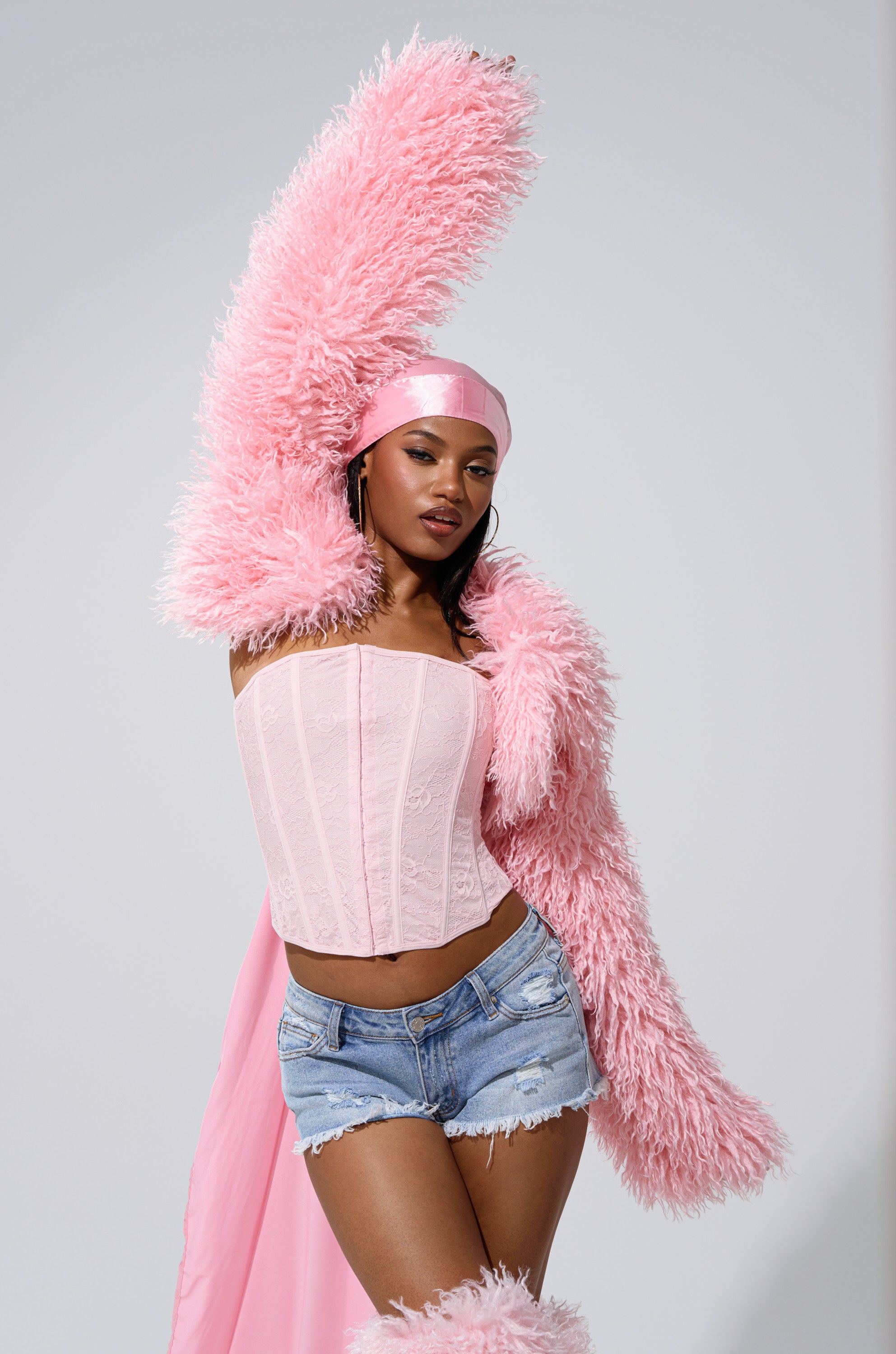 A woman poses confidently against a plain background, draping the LEILA ULTRA CROP FAUX FUR IN LIGHT PINK over her arm, paired with a pink corset top, distressed denim shorts, and a satin headscarf.