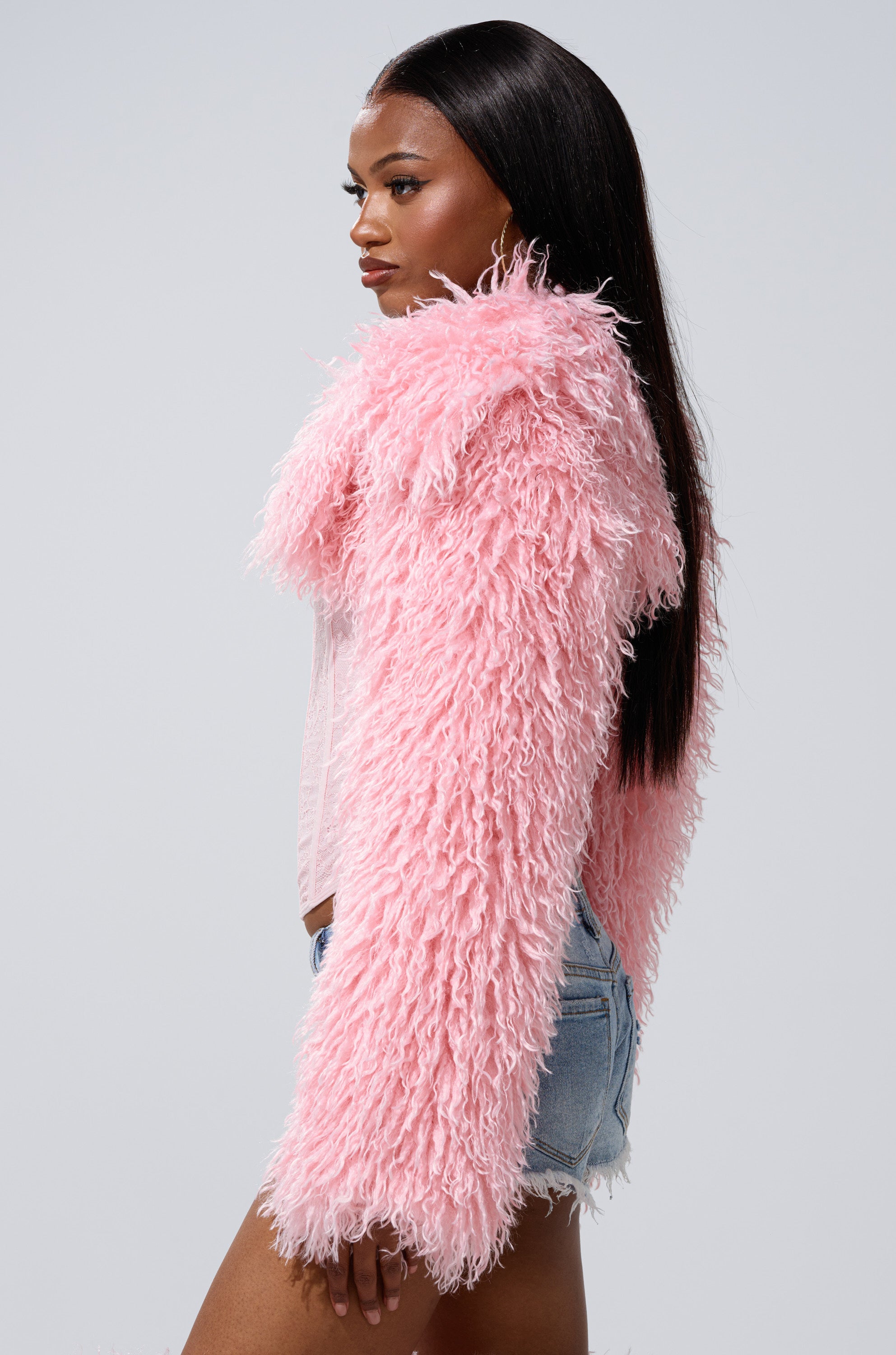 A woman with long straight hair, wearing the LEILA ULTRA CROP FAUX FUR IN LIGHT PINK and denim shorts, stands in profile against a light gray background.