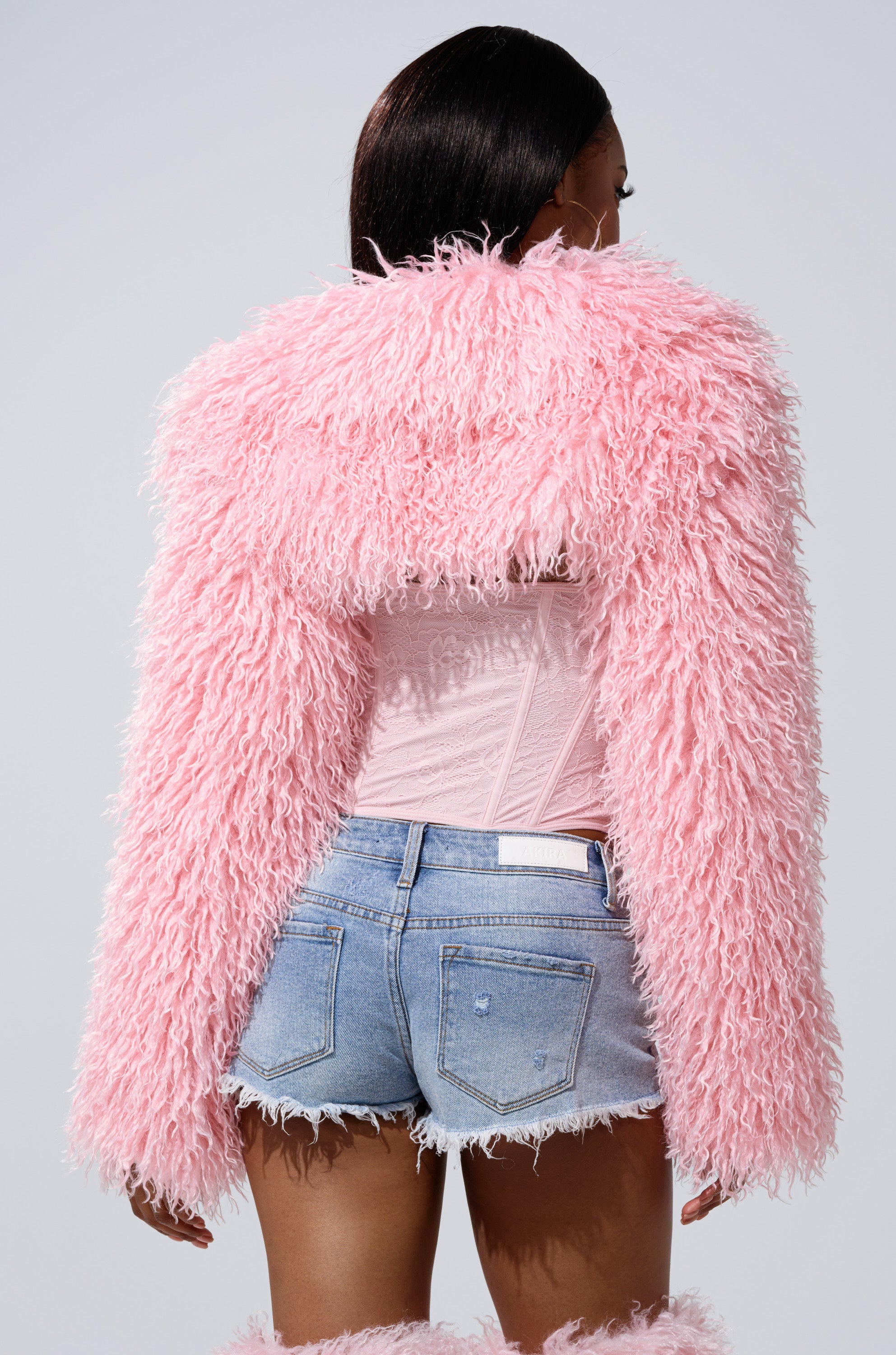 A person faces away against a plain light gray background, wearing the LEILA ULTRA CROP FAUX FUR IN LIGHT PINK jacket, a sheer pink corset top, and distressed denim shorts.