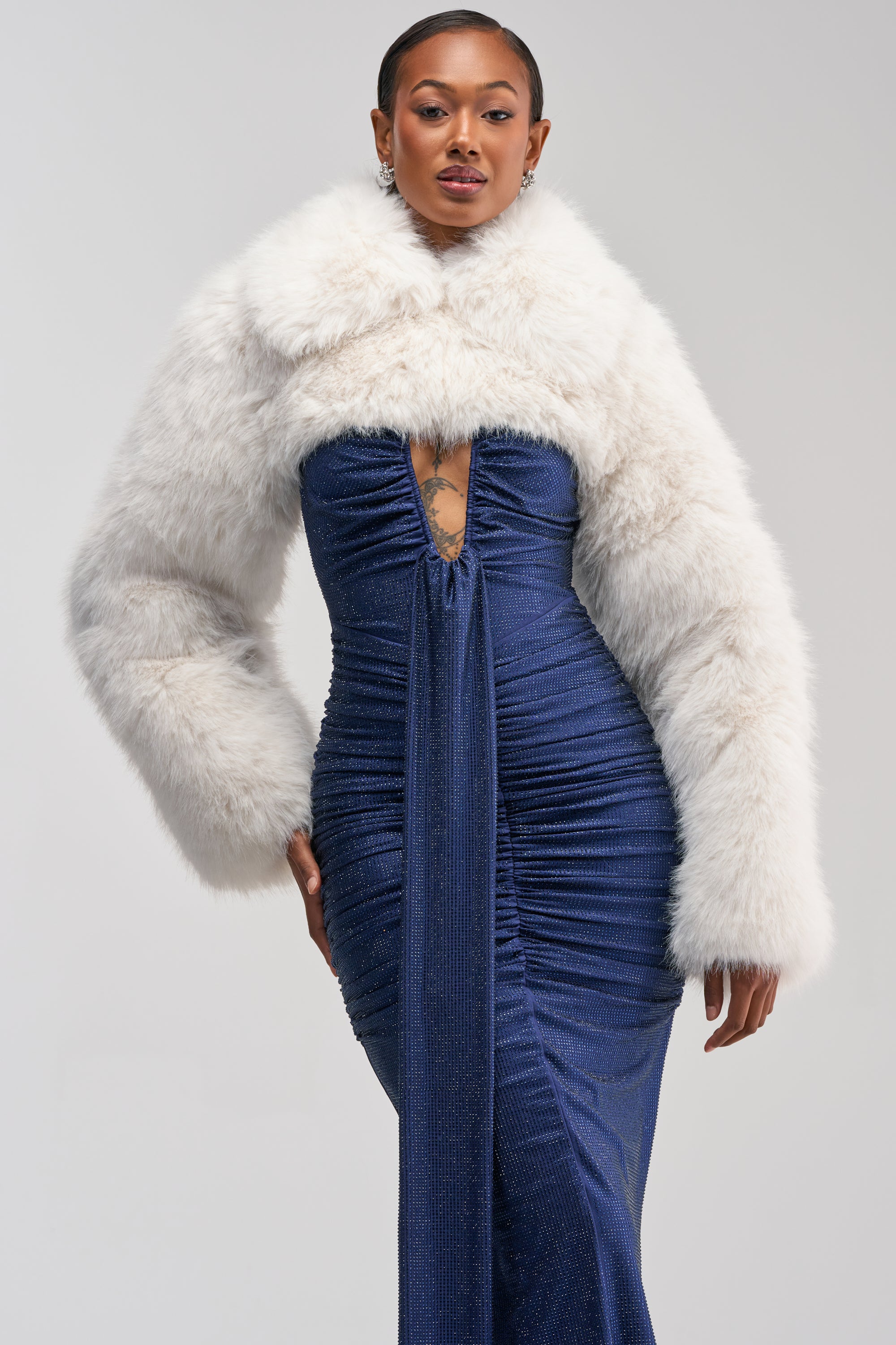A woman in a blue ruched gown with a deep neckline poses confidently, wearing the LEILA ULTRA CROP FAUX FUR IN IVORY—an elegant evening jacket perfect for luxe winter styling.