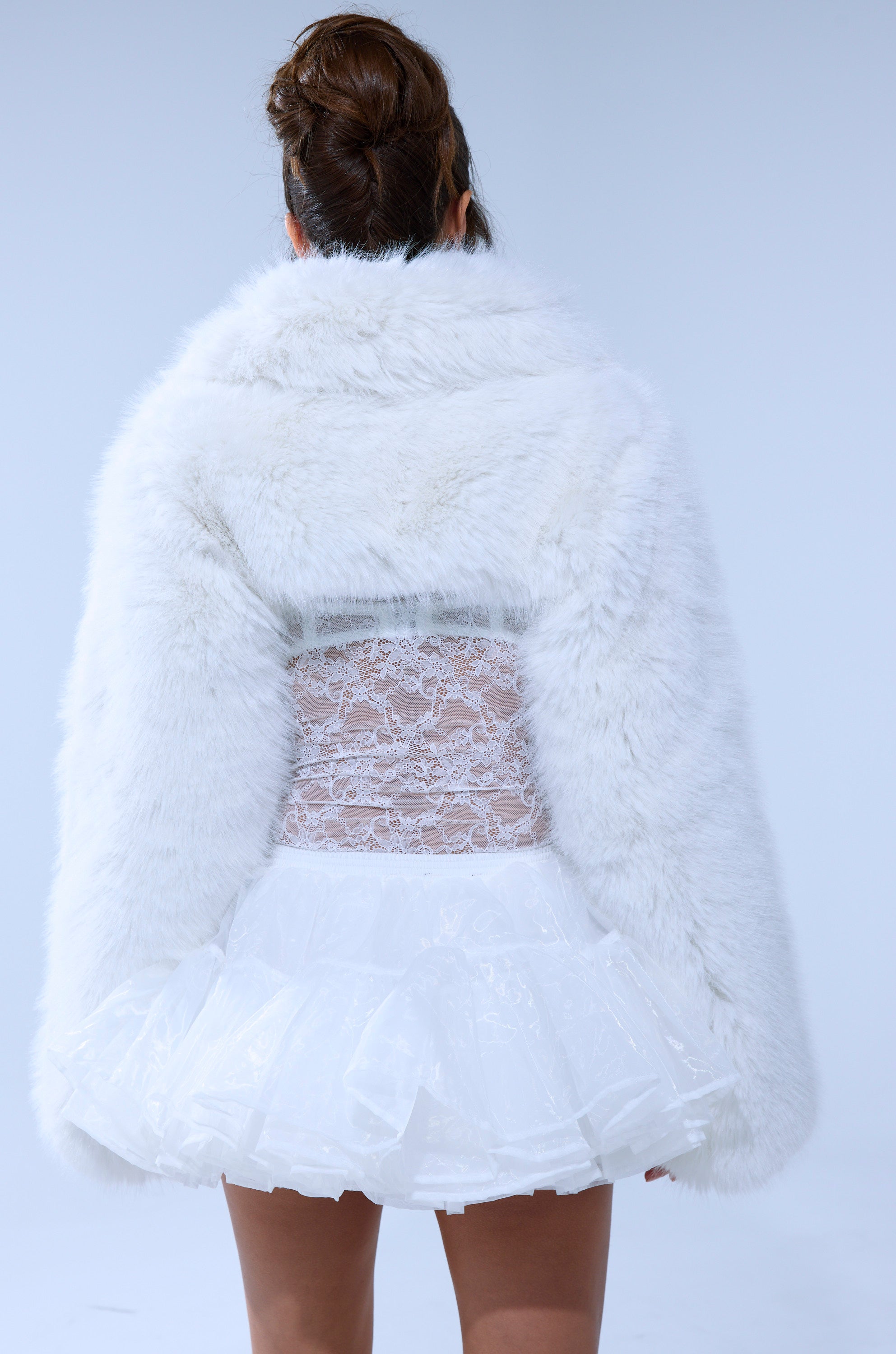A person with their back to the camera wears the LEILA ULTRA CROP FAUX FUR IN IVORY over a short, frilly white skirt and sheer lace top. Their hair is styled in a high bun against a plain light background.