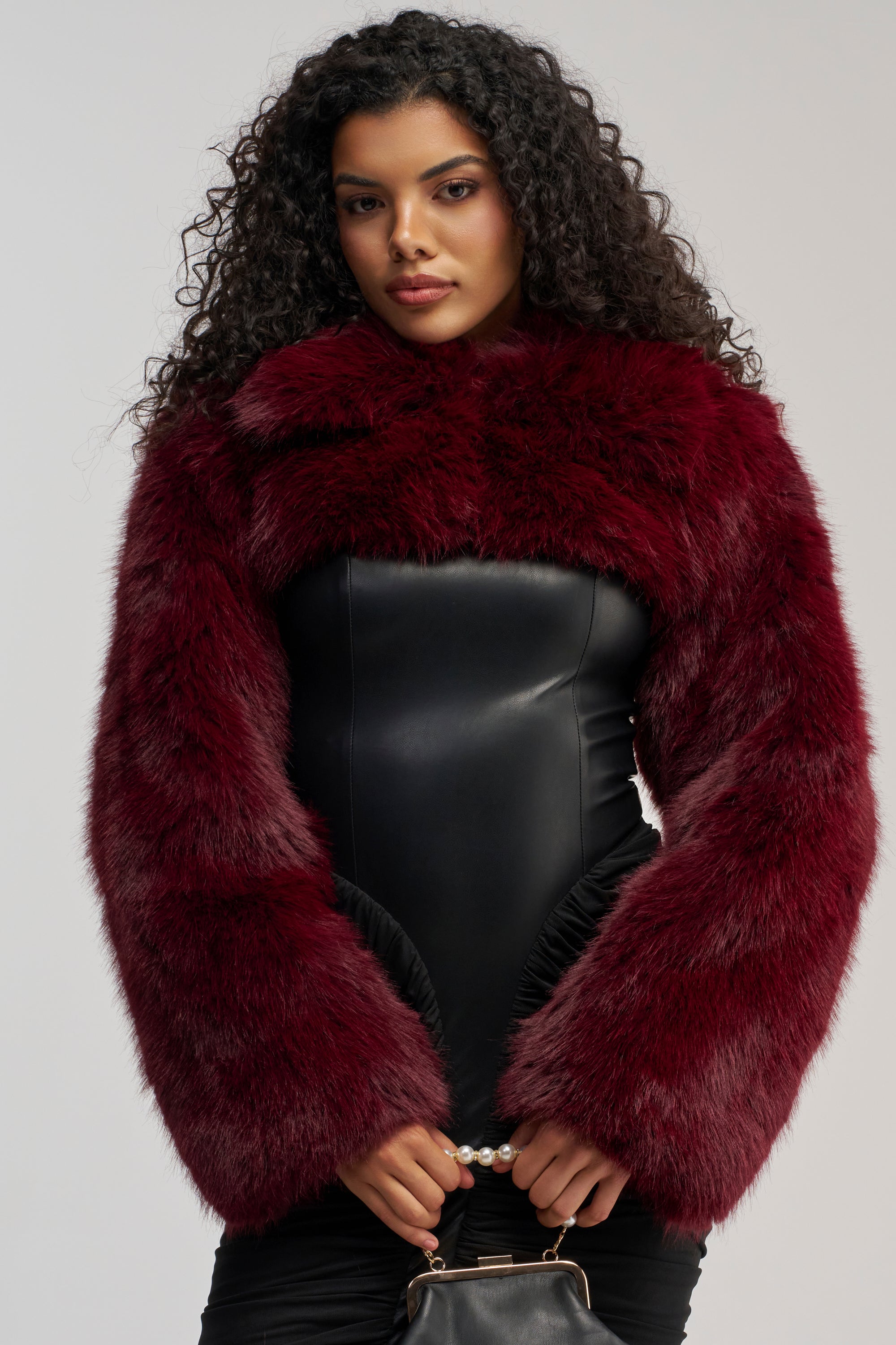 A woman with curly hair wears the LEILA ULTRA CROP FAUX FUR IN BURGUNDY jacket over a black fitted dress, holding a small black handbag with both hands, standing against a plain light background.