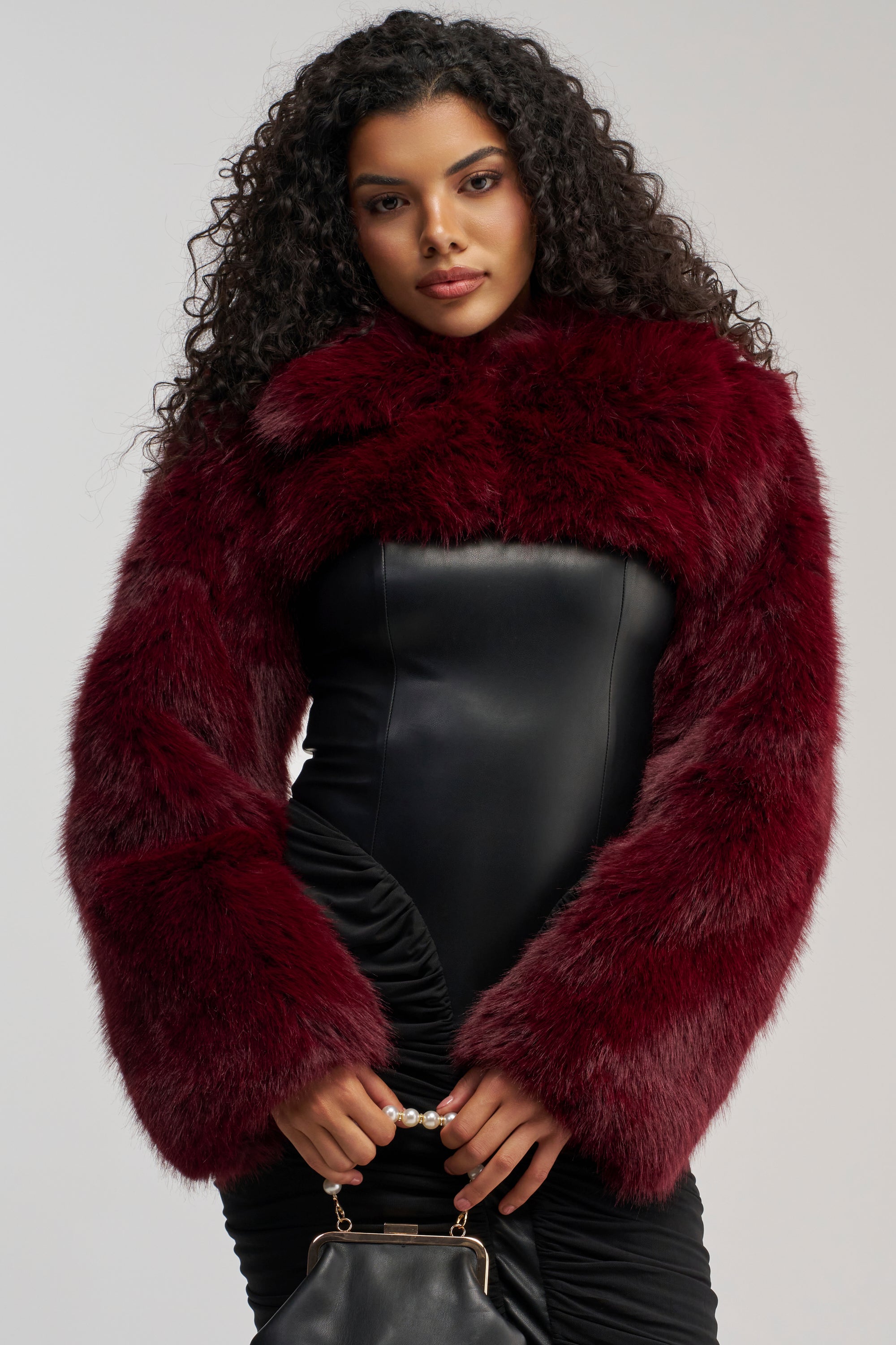 A woman with curly hair wears the LEILA ULTRA CROP FAUX FUR IN BURGUNDY over a black strapless dress and holds a small black handbag with a pearl handle, standing against a plain light background.