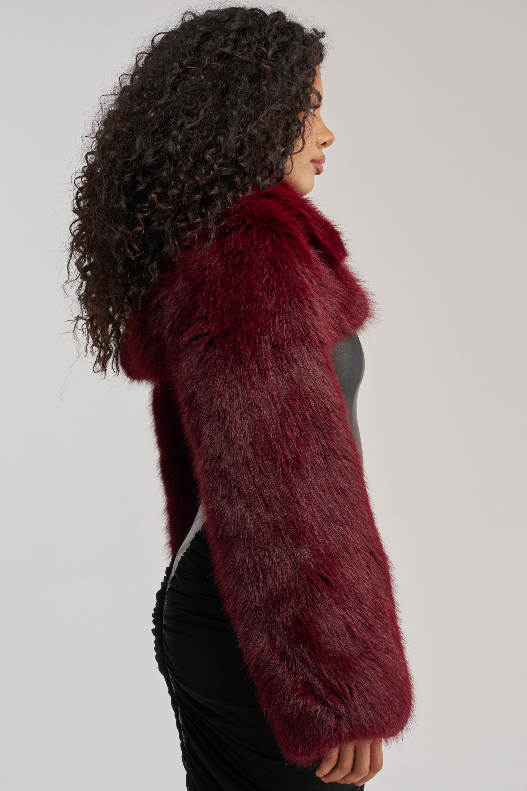 Wearing the LEILA ULTRA CROP FAUX FUR IN BURGUNDY over a black ruched dress, a person with long curly hair stands in profile against a plain light background.