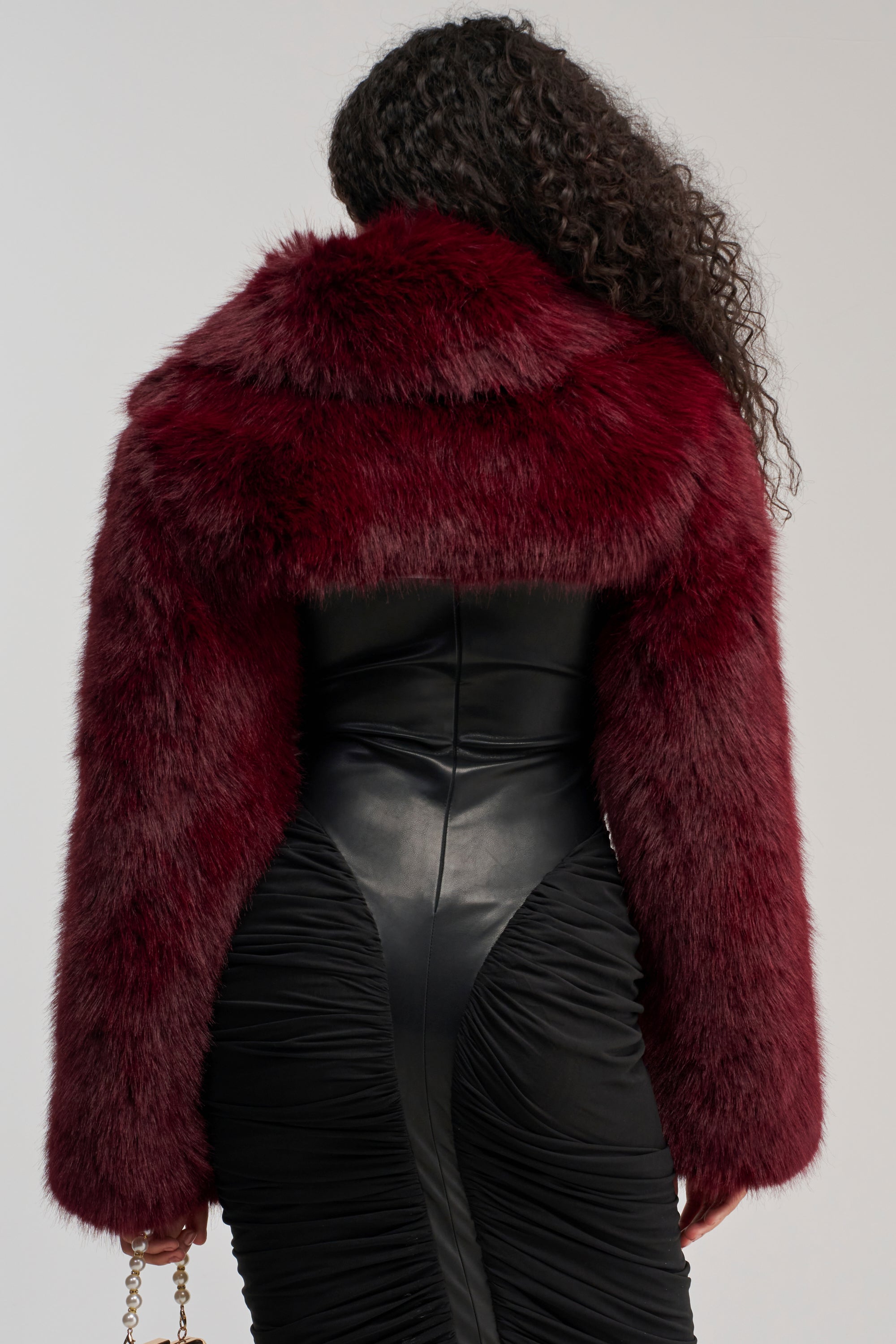 A person with long curly hair is seen from the back against a plain background, wearing the LEILA ULTRA CROP FAUX FUR IN BURGUNDY over a fitted black dress.