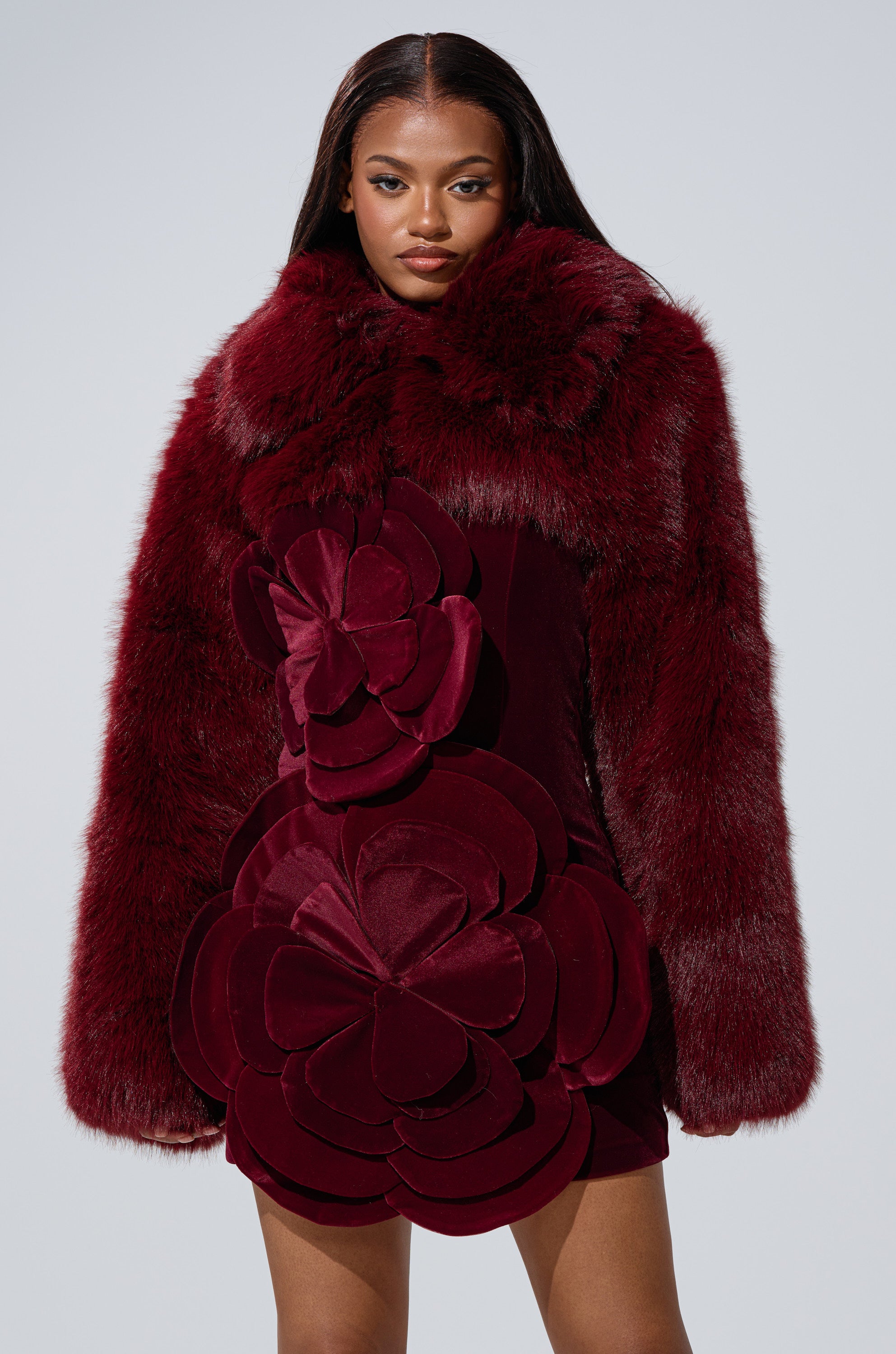 A woman models the LEILA ULTRA CROP FAUX FUR IN BURGUNDY, featuring large matching velvet floral designs, against a plain light gray background.