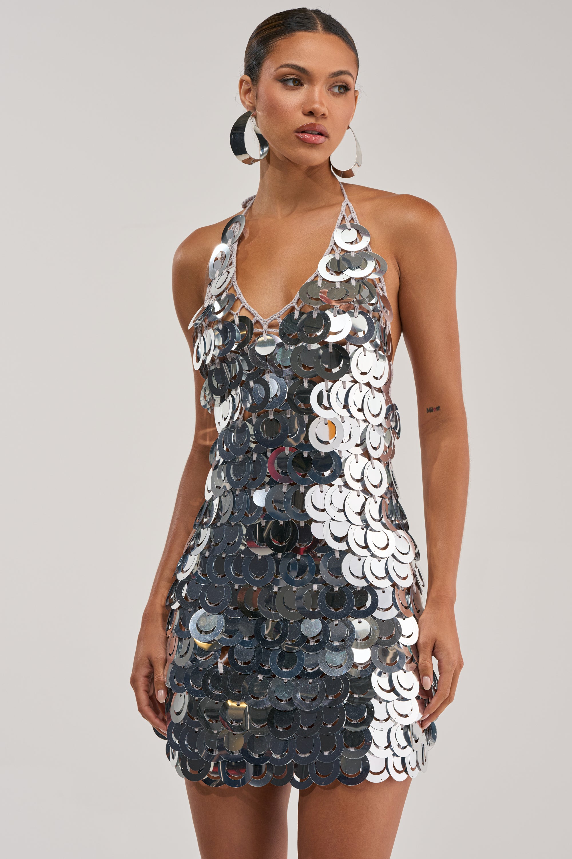 A woman wears the SHIMMER AND SHAKE SEQUIN MINI DRESS IN SILVER, a sleeveless, deep V-neck halter mini dress with large shiny sequins. Paired with hoop earrings and slicked-back hair, it's perfect for bold festival looks against a simple backdrop.