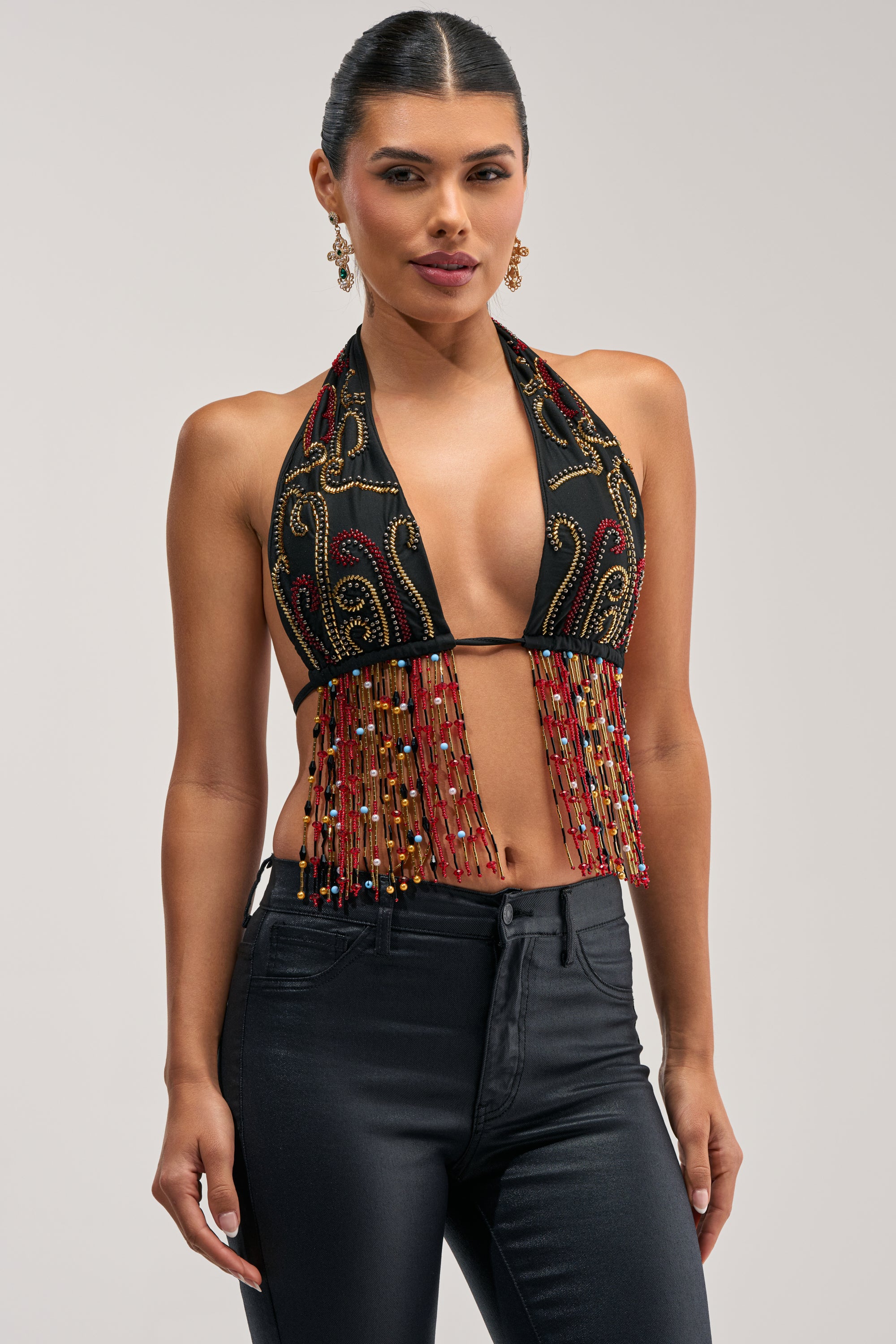 A woman with her hair pulled back models the ANGEL ON EARTH BEADED BRALETTE TOP, styled as a halter featuring black fabric with gold embroidery and dangling red, gold, and light blue beaded fringe. She pairs the festival bralette with black pants against a plain, light gray background.