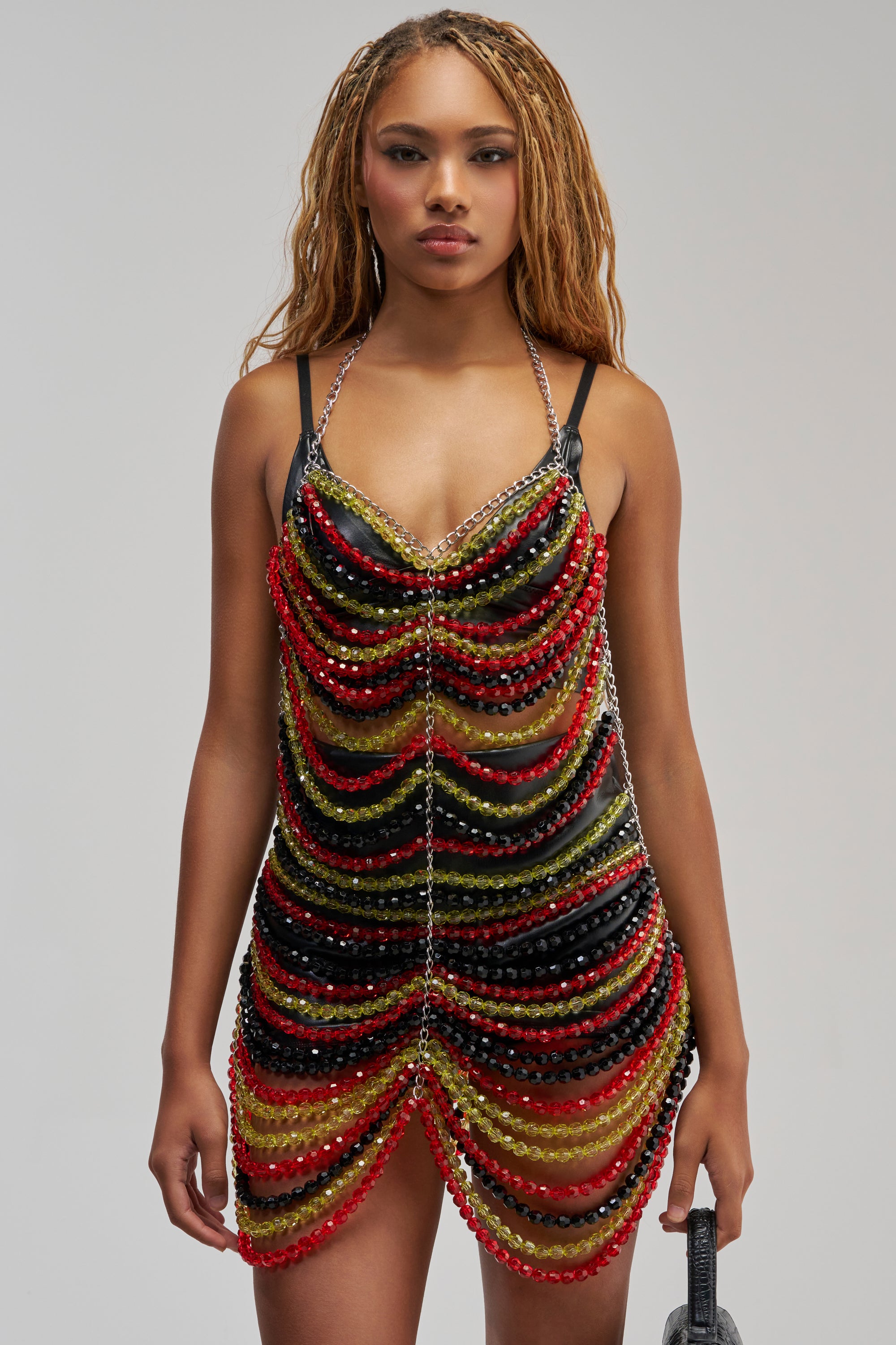 A woman with long, braided hair wears the CARNIVALE BEADED MINI DRESS IN GREEN MULTI—a sleeveless festival dress made of layered green, yellow, and black beads—standing against a plain gray background.