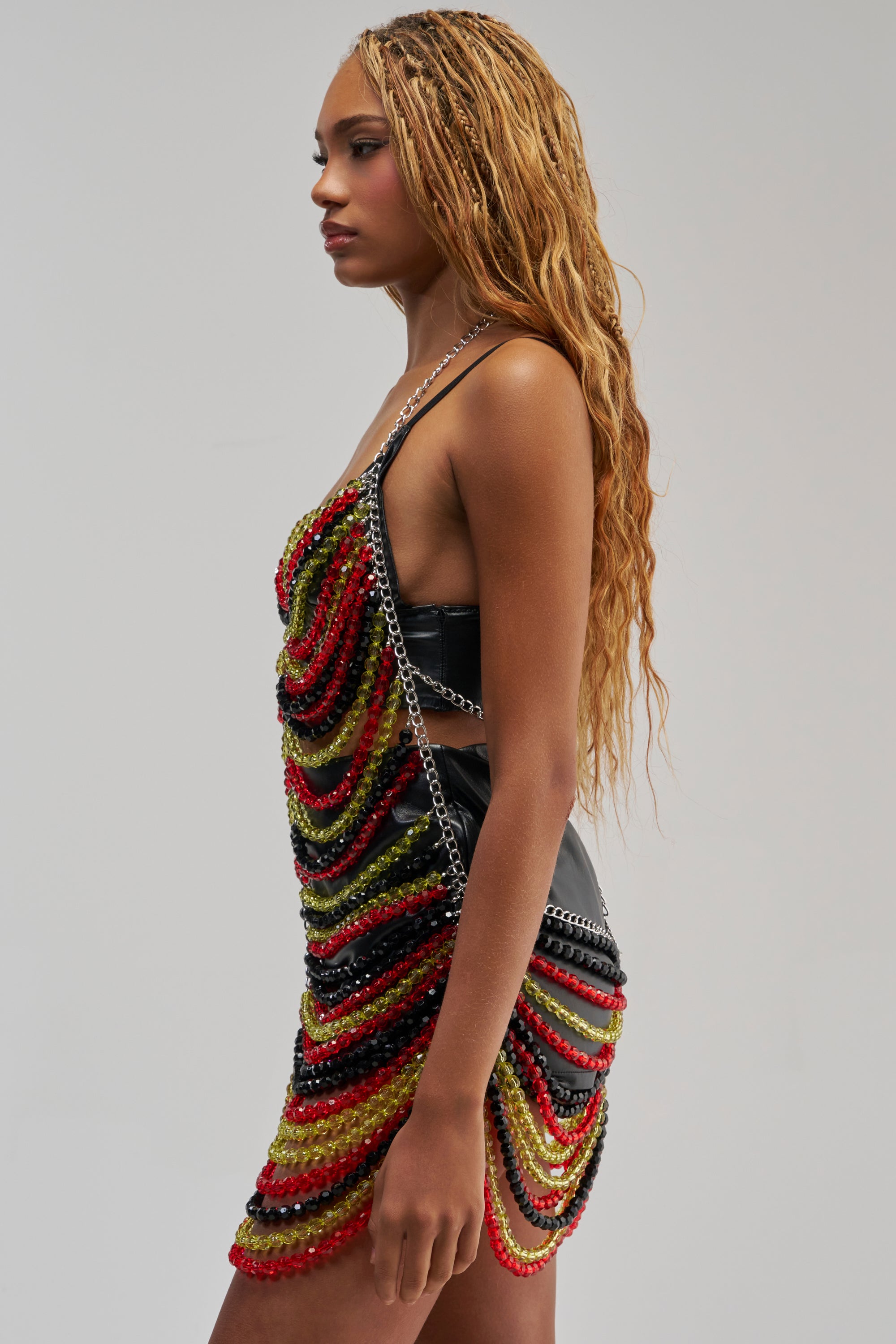A person with long, wavy hair poses in profile against a plain light background, wearing the CARNIVALE BEADED MINI DRESS IN GREEN MULTI—a festival dress featuring a black beaded chain halter silhouette adorned with layered green, yellow, and black beads.