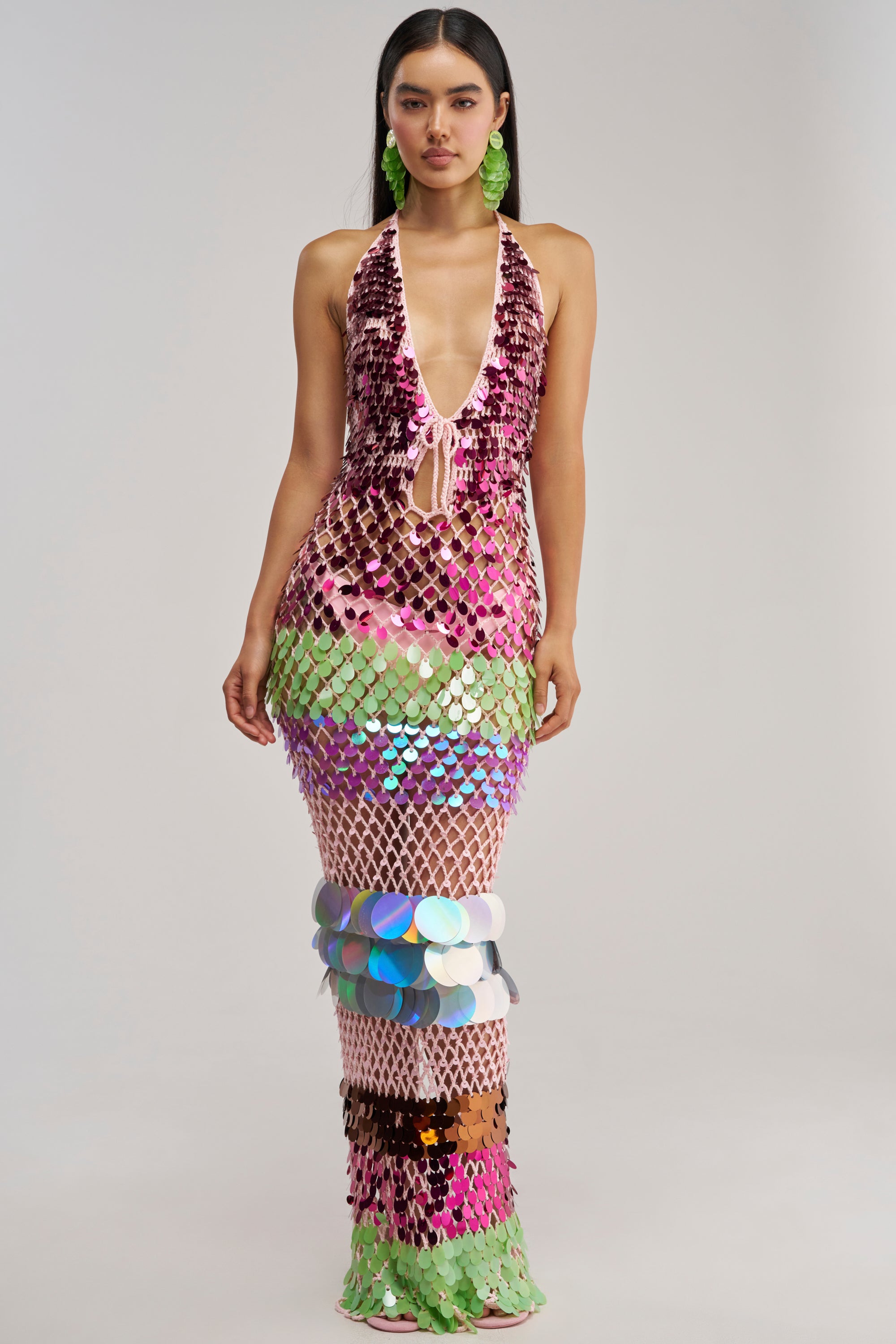 Dressed in the PARTY CRASHER SEQUIN MAXI DRESS IN GREEN MULTI—a long, form-fitting crochet halter maxi adorned with large, vibrant sequins in pink, green, silver, and holographic hues—she stuns with a deep plunging neckline. Perfectly complemented by bold green statement earrings, this show-stopping dress is ideal for a glamorous vacation night or an unforgettable beach party.