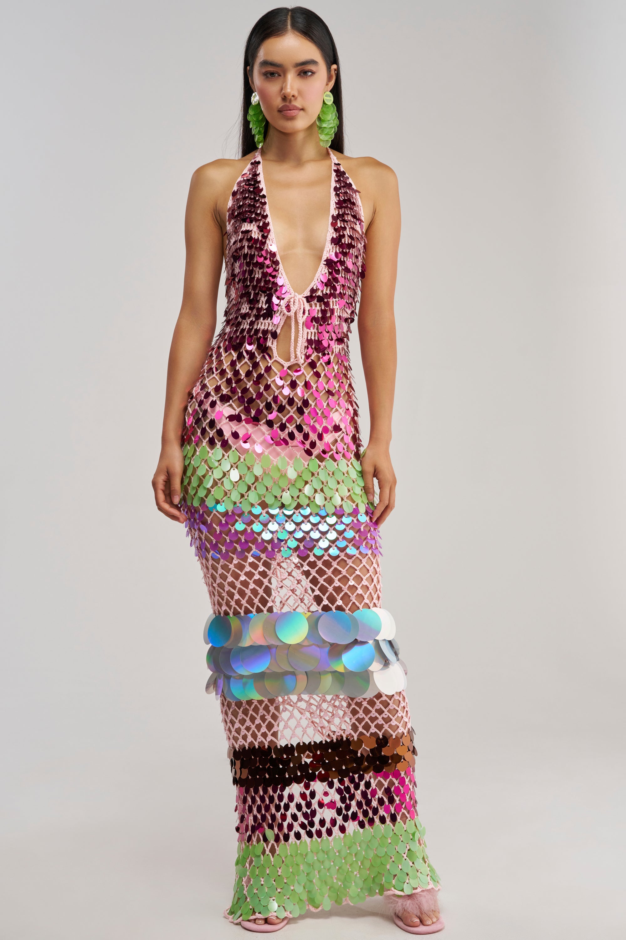 A woman stands wearing the PARTY CRASHER SEQUIN MAXI DRESS IN GREEN MULTI, a dazzling sequin crochet halter maxi dress with a sleeveless, deep V-neck and floor-length silhouette, adorned with shimmering discs in green, pink, purple, and blue. She accessorizes with long hair, green earrings, and pink sandals against a plain background—perfect as a vibrant beach party outfit or an unforgettable vacation night dress.