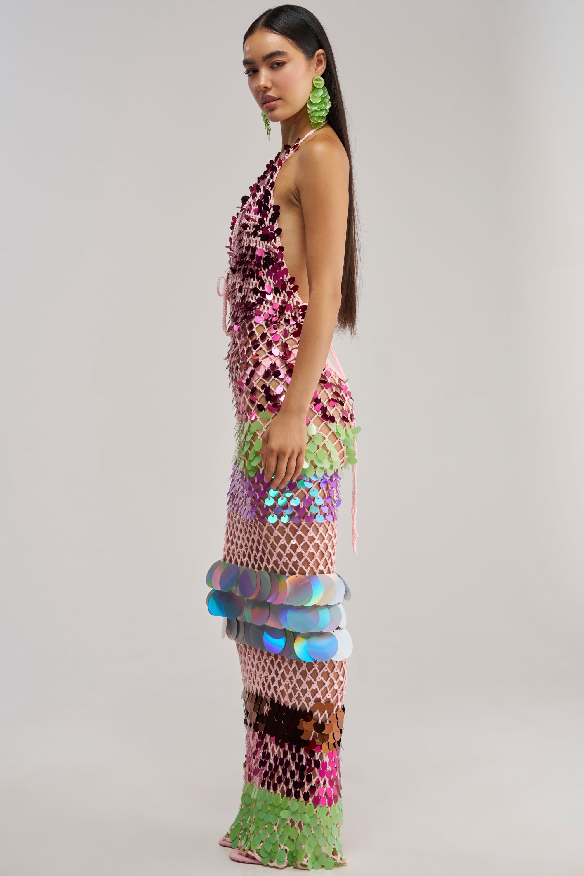 A model poses in profile wearing the PARTY CRASHER SEQUIN MAXI DRESS IN GREEN MULTI—a long, backless crochet dress adorned with large, iridescent, metallic rainbow sequins—perfect for a vacation night out or as a show-stopping dress for a beach party. She completes the look with bold, lime-green circular earrings against a plain light gray studio background.