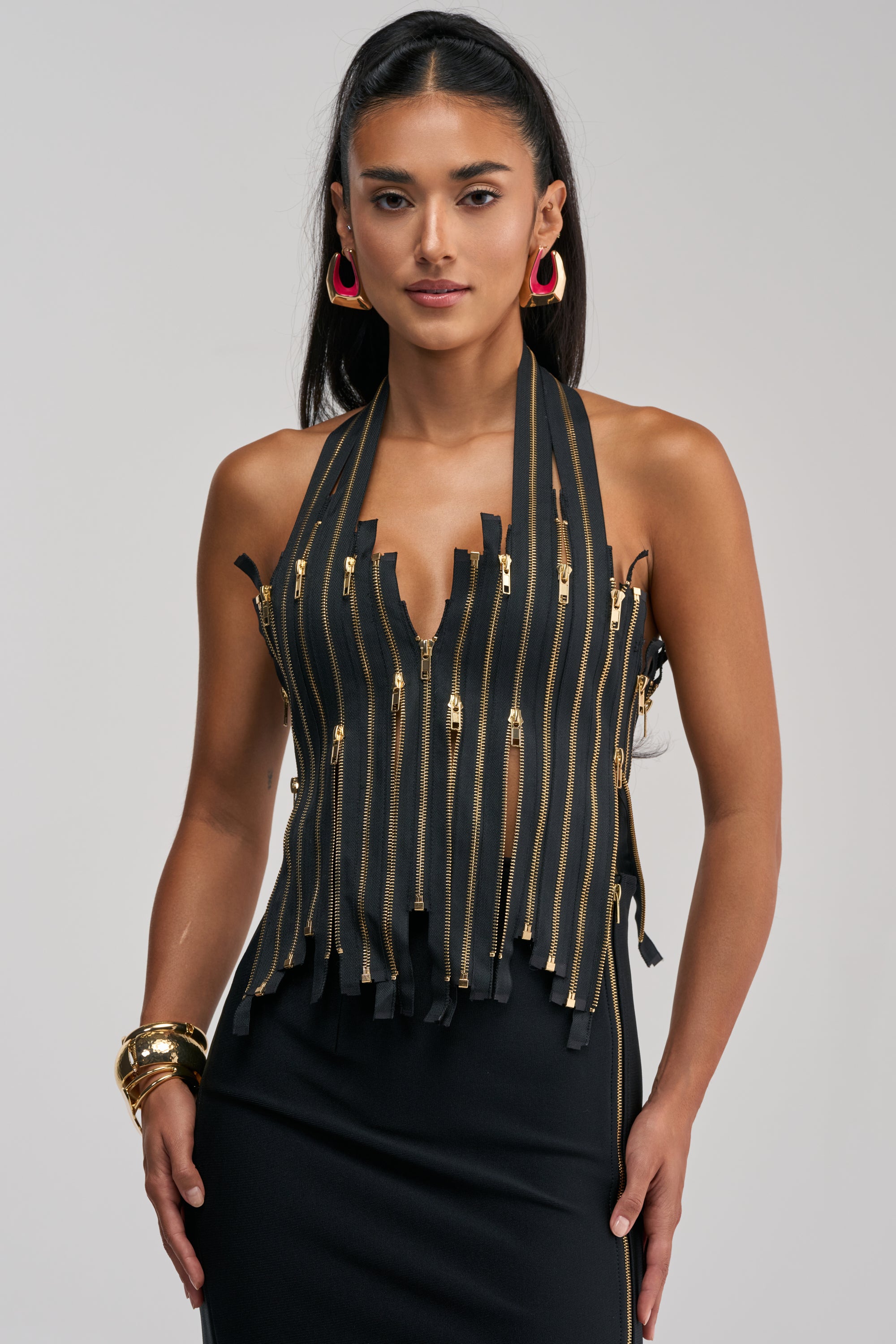 A woman is wearing the YOU REALLY THOUGHT ZIP UP CORSET—a sleek, sleeveless black top with multiple vertical zippers—paired with a black skirt, gold bangles, and pink hoop earrings as she stands against a plain background.