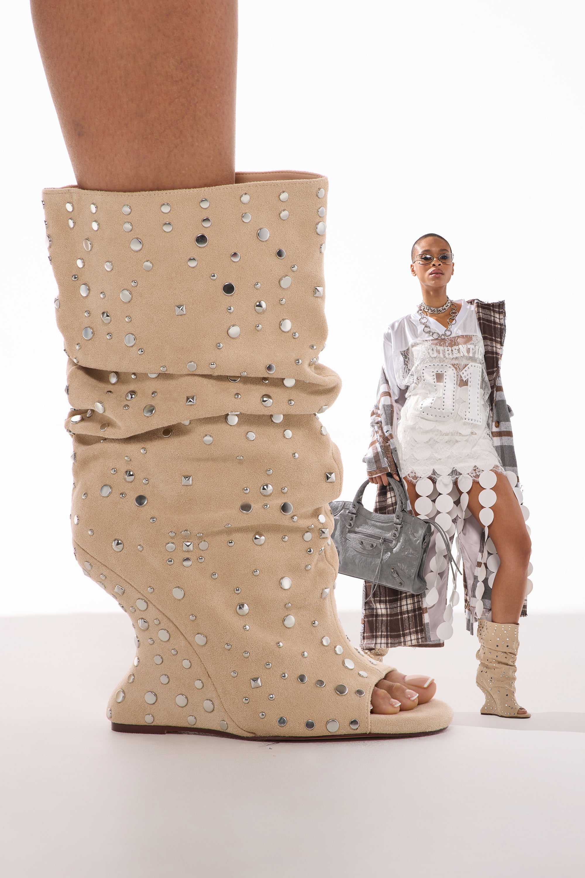 A close-up of the AZALEA WANG NAOMANI FAUX SUEDE BEIGE STUDDED OPEN TOE HEEL, with silver studs, as a stylish person in white and sunglasses holds a gray bag—also wearing these boho-chic heels. 1