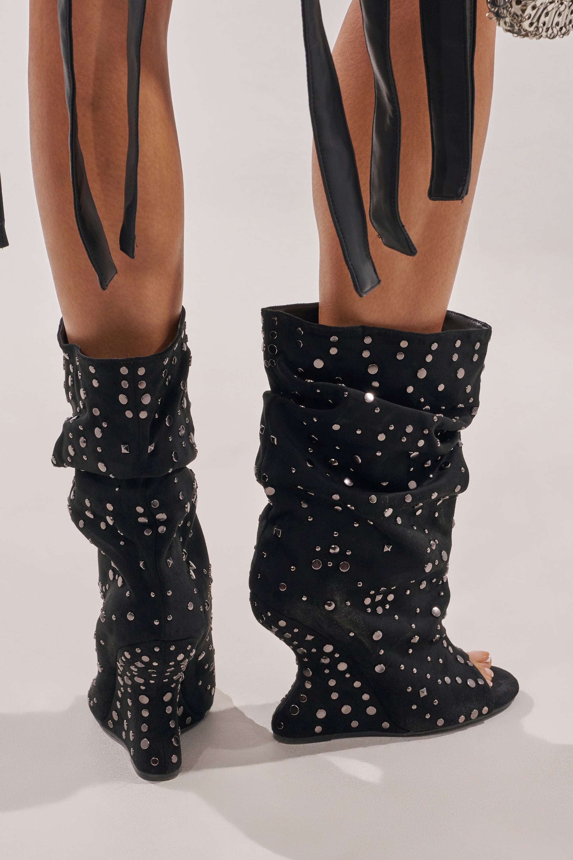A person stands on a light surface wearing the AZALEA WANG NAOMANI FAUX SUEDE BLACK STUDDED OPEN TOE BOOTIE—black, slouchy wedge booties with silver studs. Black ribbon-like strips hang from the upper part of their outfit. 2