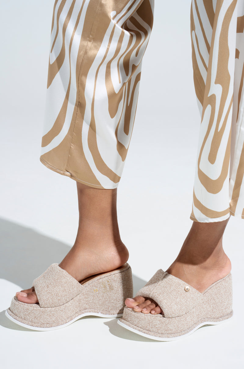 The individual is stylishly adorned in the AZALEA WANG NATHALIA Beige Novelty Flatform Sandals, featuring a platform sole and gold button accents, paired with cream and brown patterned wide-leg pants. The textured fabric pops beautifully against a light neutral backdrop.