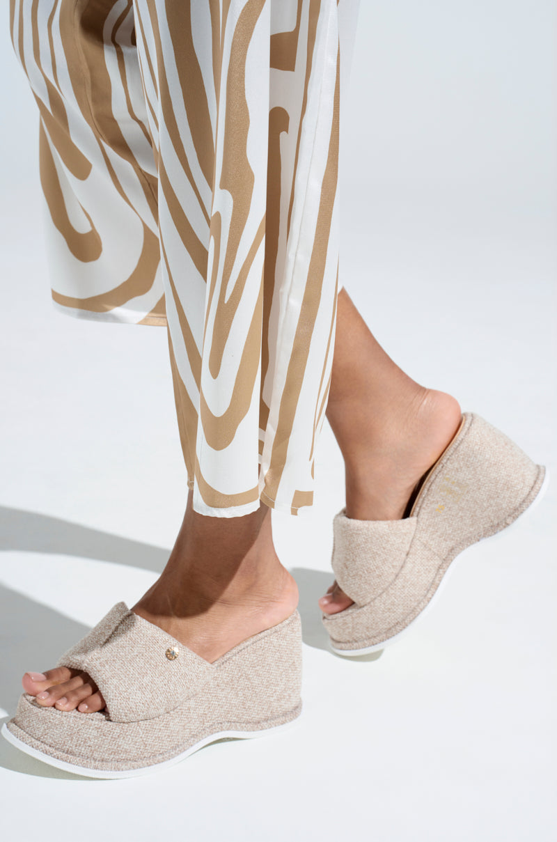A close-up shows a person wearing AZALEA WANG NATHALIA BEIGE NOVELTY FLATFORM SANDALS with open toes and cushioned insoles. The textured fabric pairs elegantly with white soles, complemented by wide-legged pants in a white and tan geometric pattern on a plain white background.