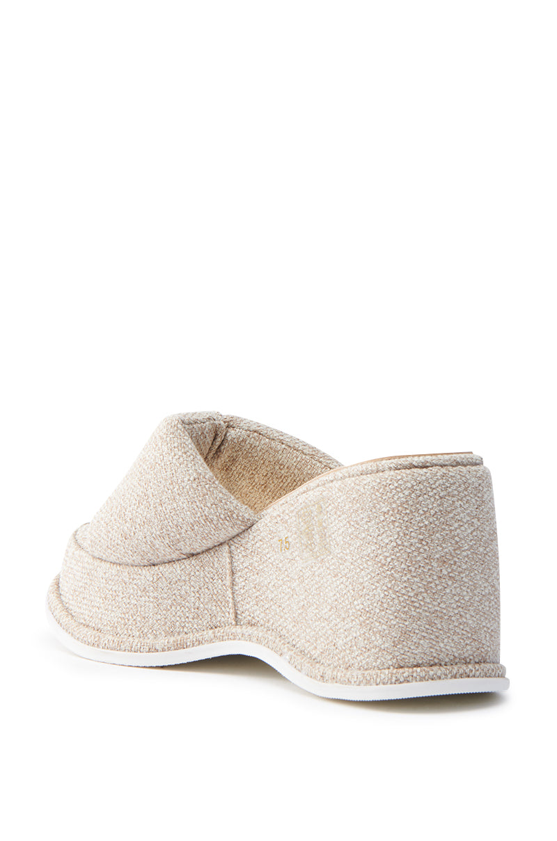 The AZALEA WANG NATHALIA beige novelty flatform sandal, seen from behind at an angle, showcases a textured fabric surface, a low back design, and a cushioned insole for comfort on a sleek white sole.