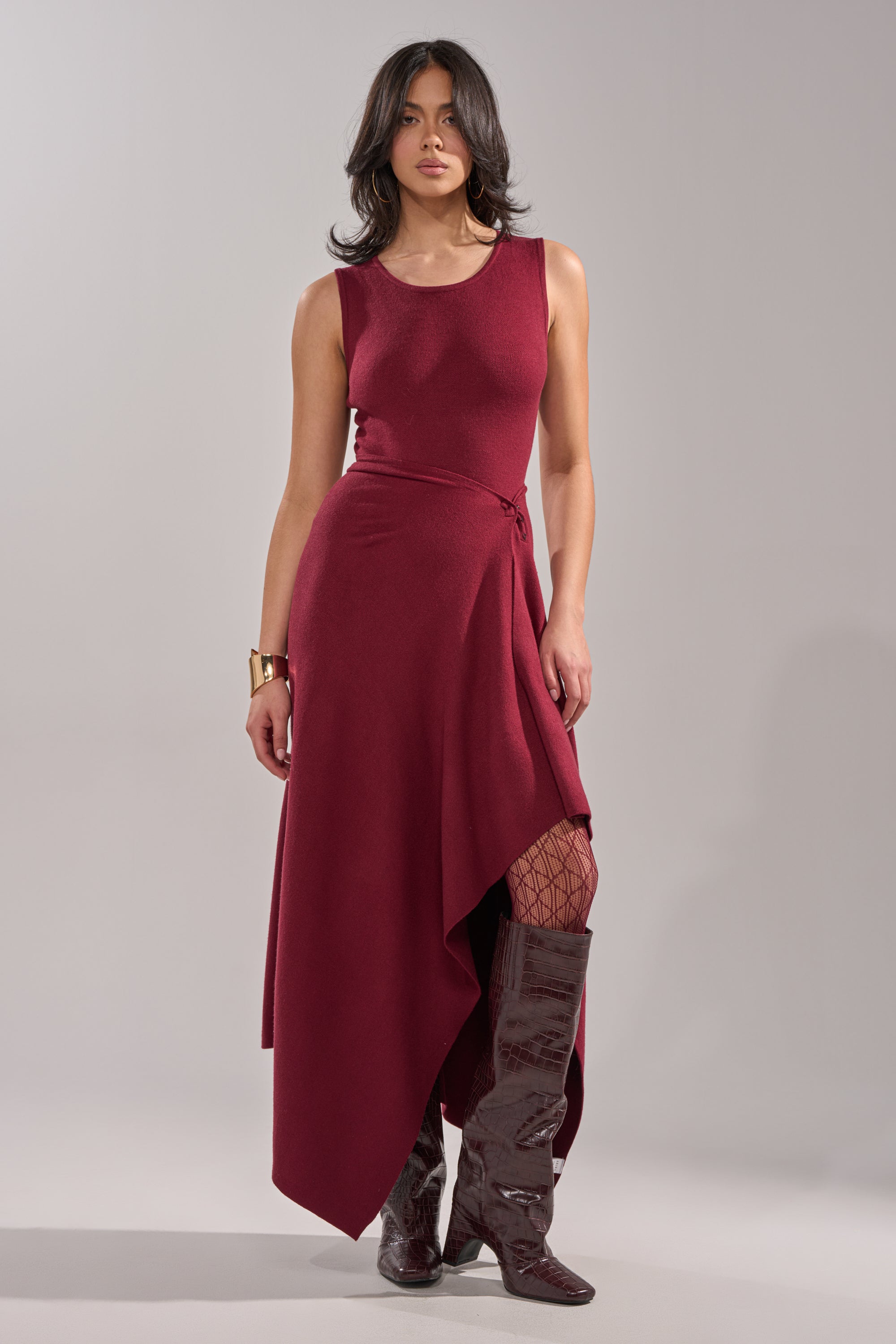 A woman poses against a plain light gray background, wearing the LOVER SWEATER MAXI SKIRT with a sleeveless top, patterned fishnet tights, glossy knee-high boots, shoulder-length dark hair, and minimal jewelry. 1
