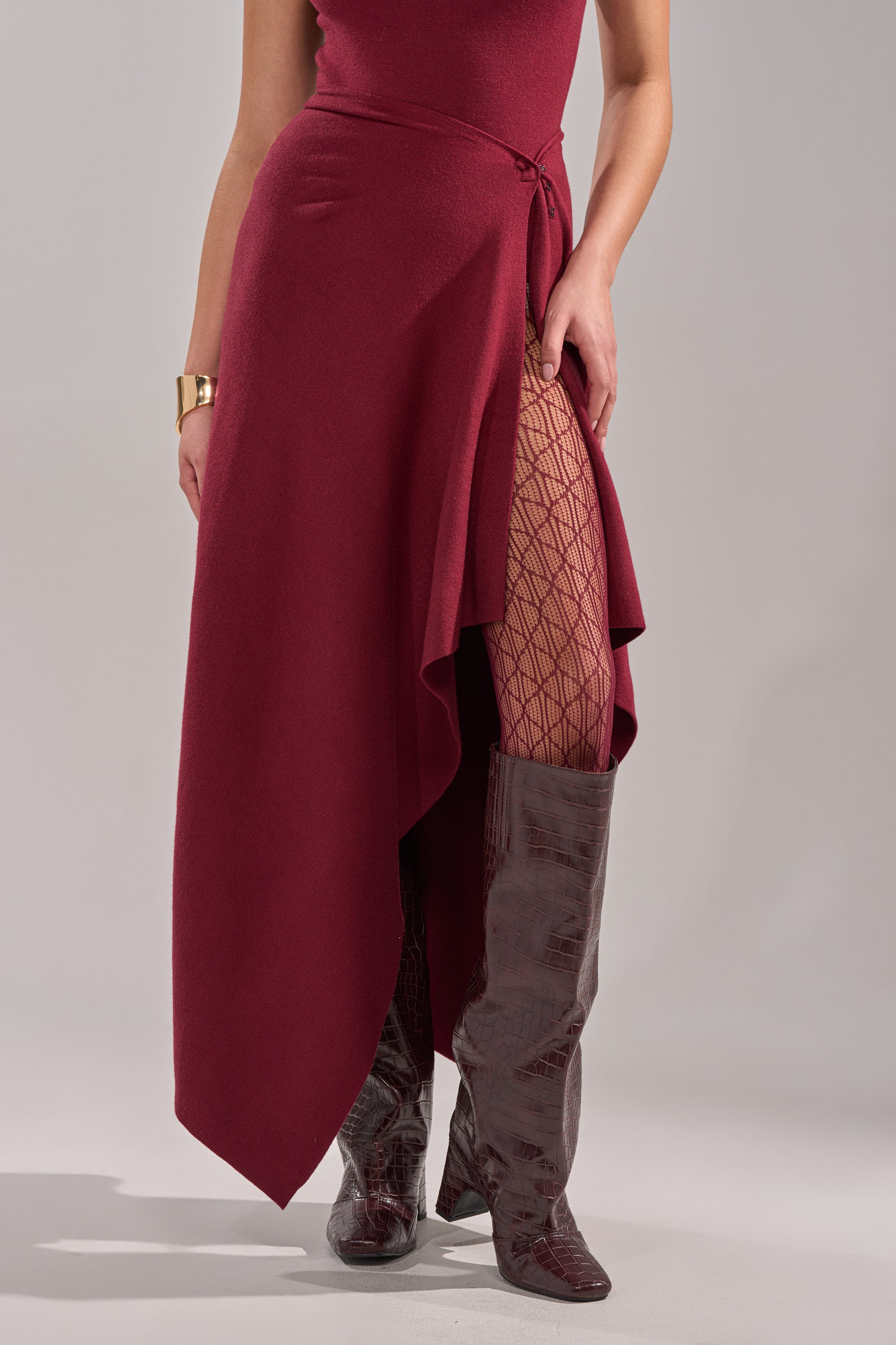 Wearing the LOVER SWEATER MAXI SKIRT in burgundy, geometric-patterned tights, textured knee-high boots, and a gold bracelet, a person stands against a neutral background. 0