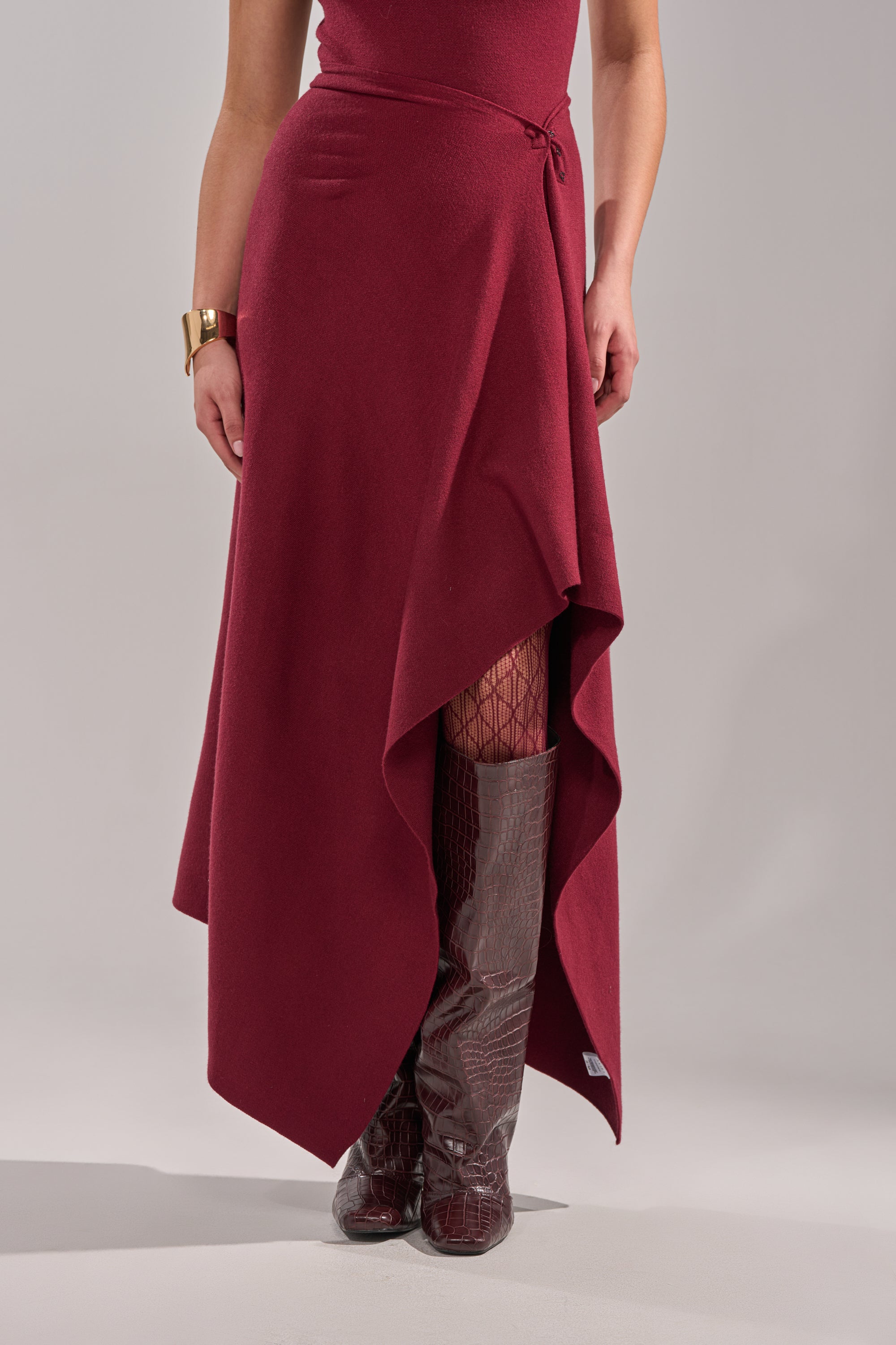 A person models the LOVER SWEATER MAXI SKIRT in wine red with dark patterned tights, shiny brown knee-high boots, and a gold bracelet, standing against a plain light background. 2