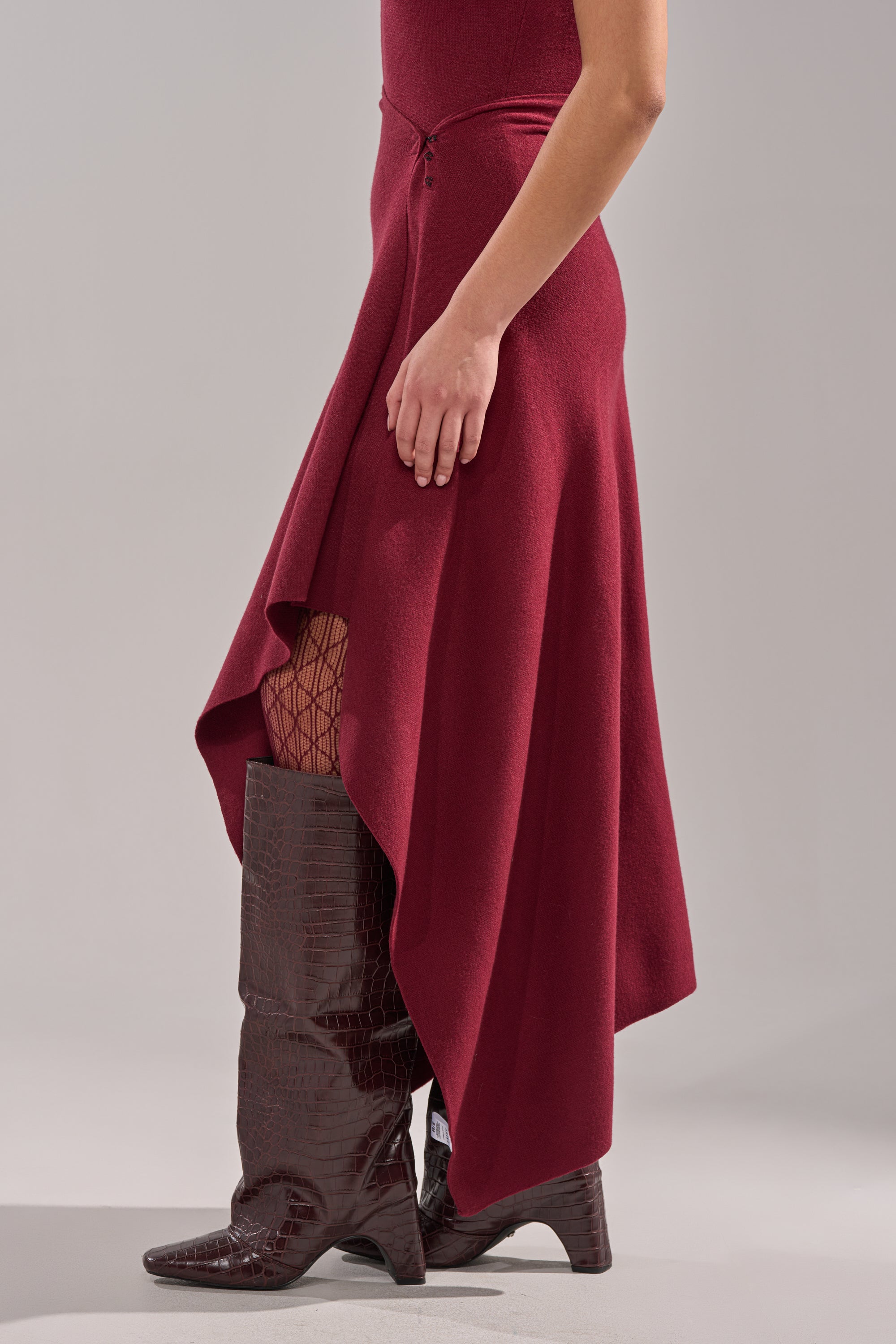 A person wearing the LOVER SWEATER MAXI SKIRT in burgundy, paired with fishnet stockings and tall dark brown crocodile-texture boots, stands against a plain light background. 3
