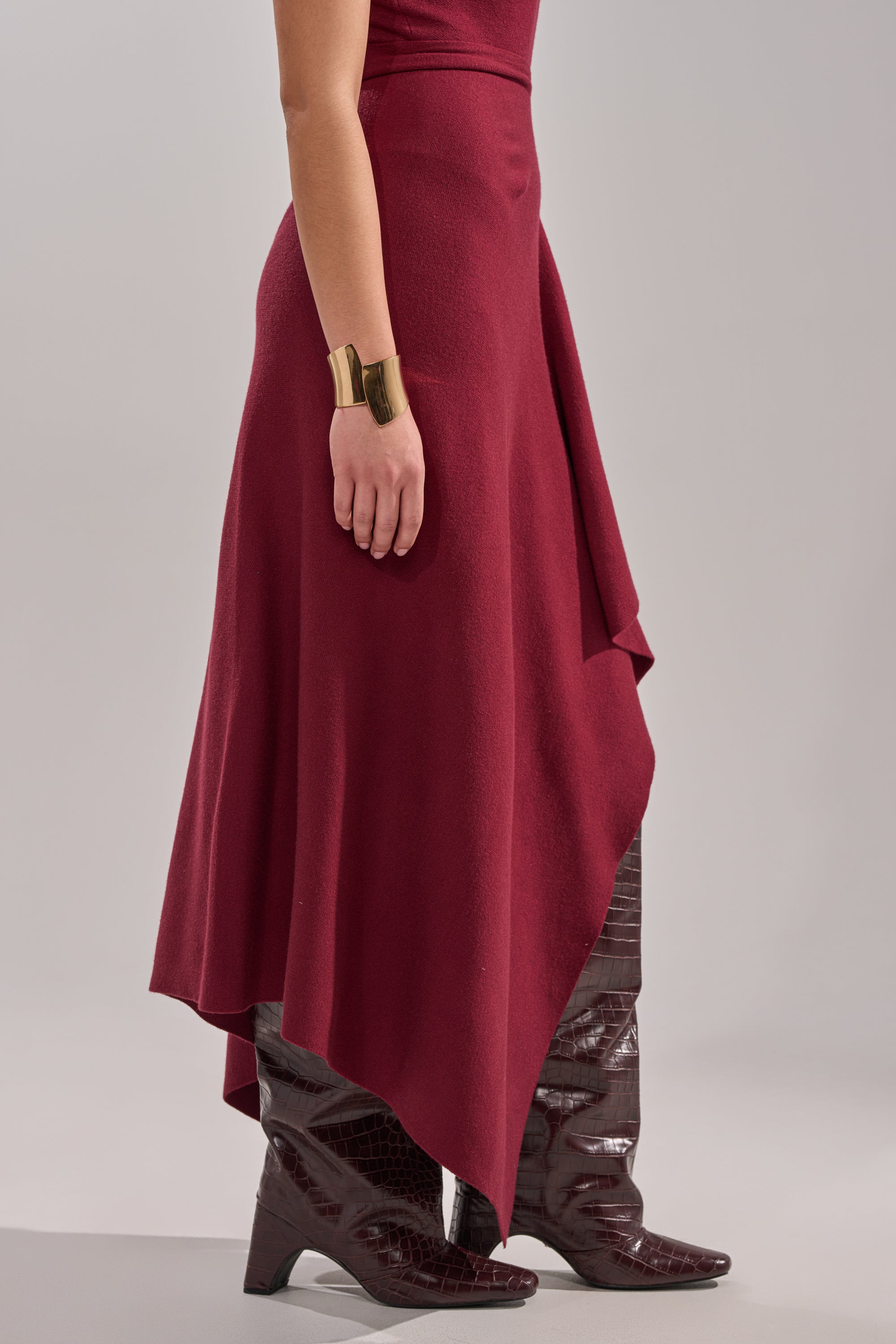 A person wears the LOVER SWEATER MAXI SKIRT styled as a sleeveless dress, paired with dark knee-high crocodile boots and a wide gold cuff bracelet, standing against a plain gray background. 4