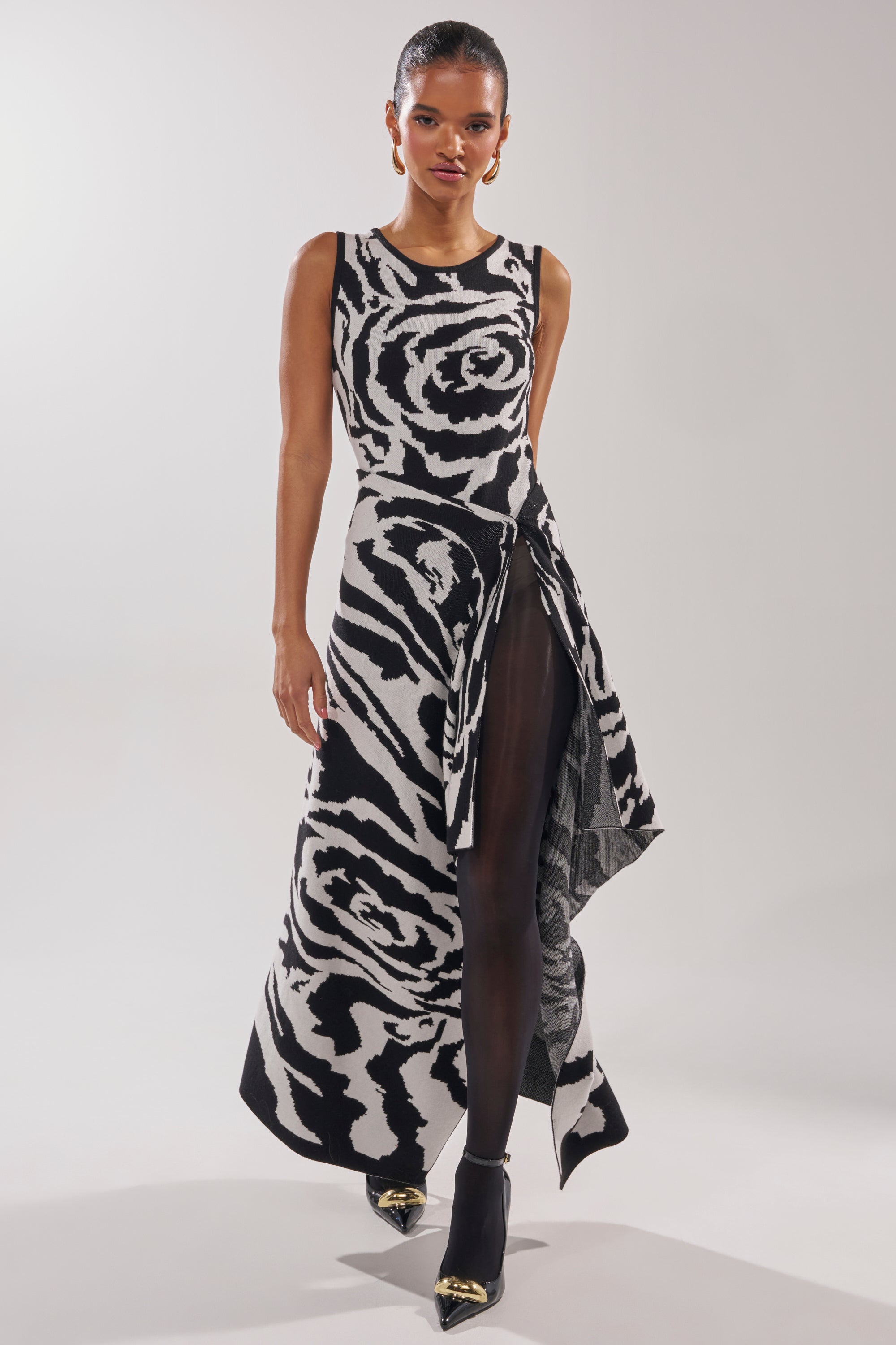 A woman models the NATOMA FLORAL MAXI SKIRT featuring bold black and white rose and zebra-like prints with an asymmetrical hem, styled with black sheer tights and pointed black heels with gold tips against a plain background.
