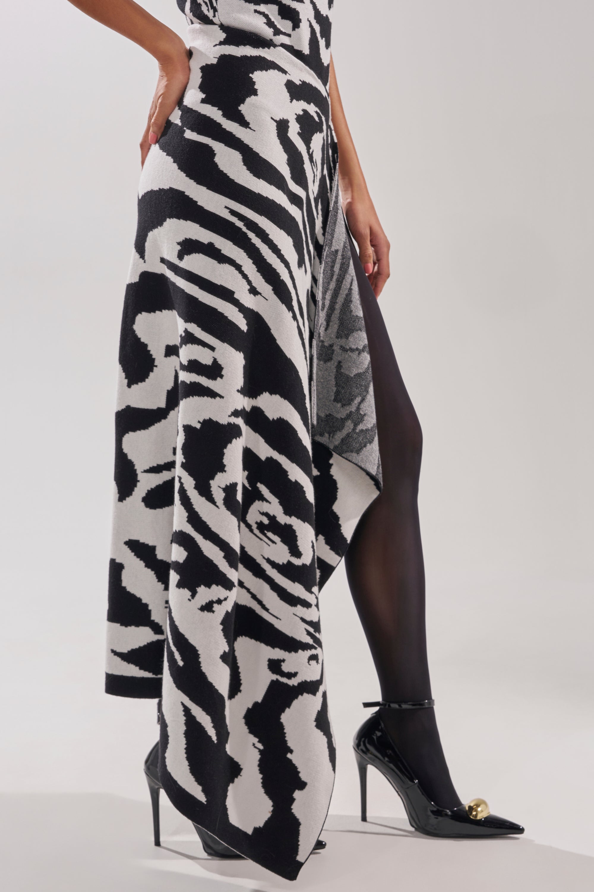 A person wearing the NATOMA FLORAL MAXI SKIRT, styled with black sheer tights and black high heels with gold embellishments, stands against a plain background.