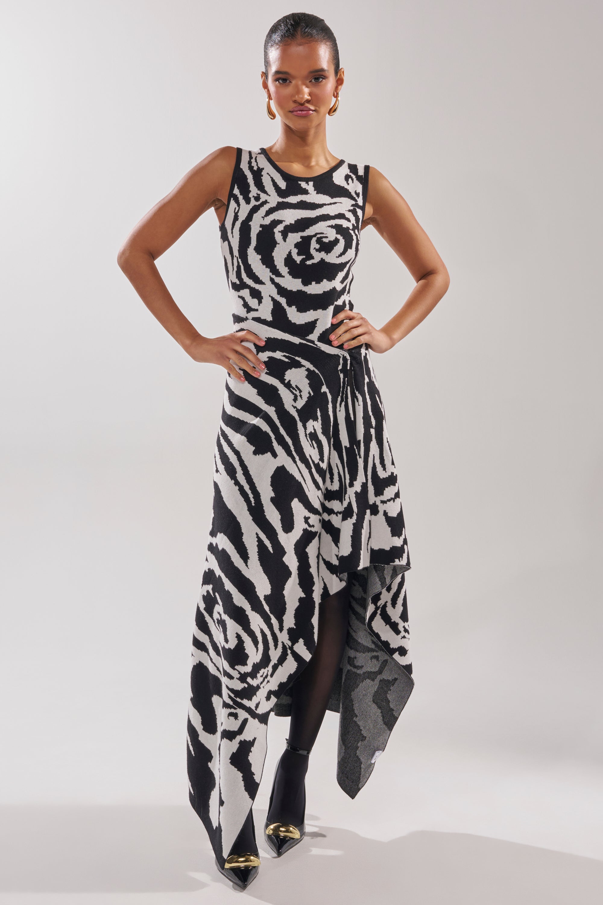 A woman stands confidently against a light background, wearing the NATOMA FLORAL MAXI SKIRT with bold black and white abstract rose patterns, paired with black ankle boots featuring metallic gold toes and her hair pulled back.