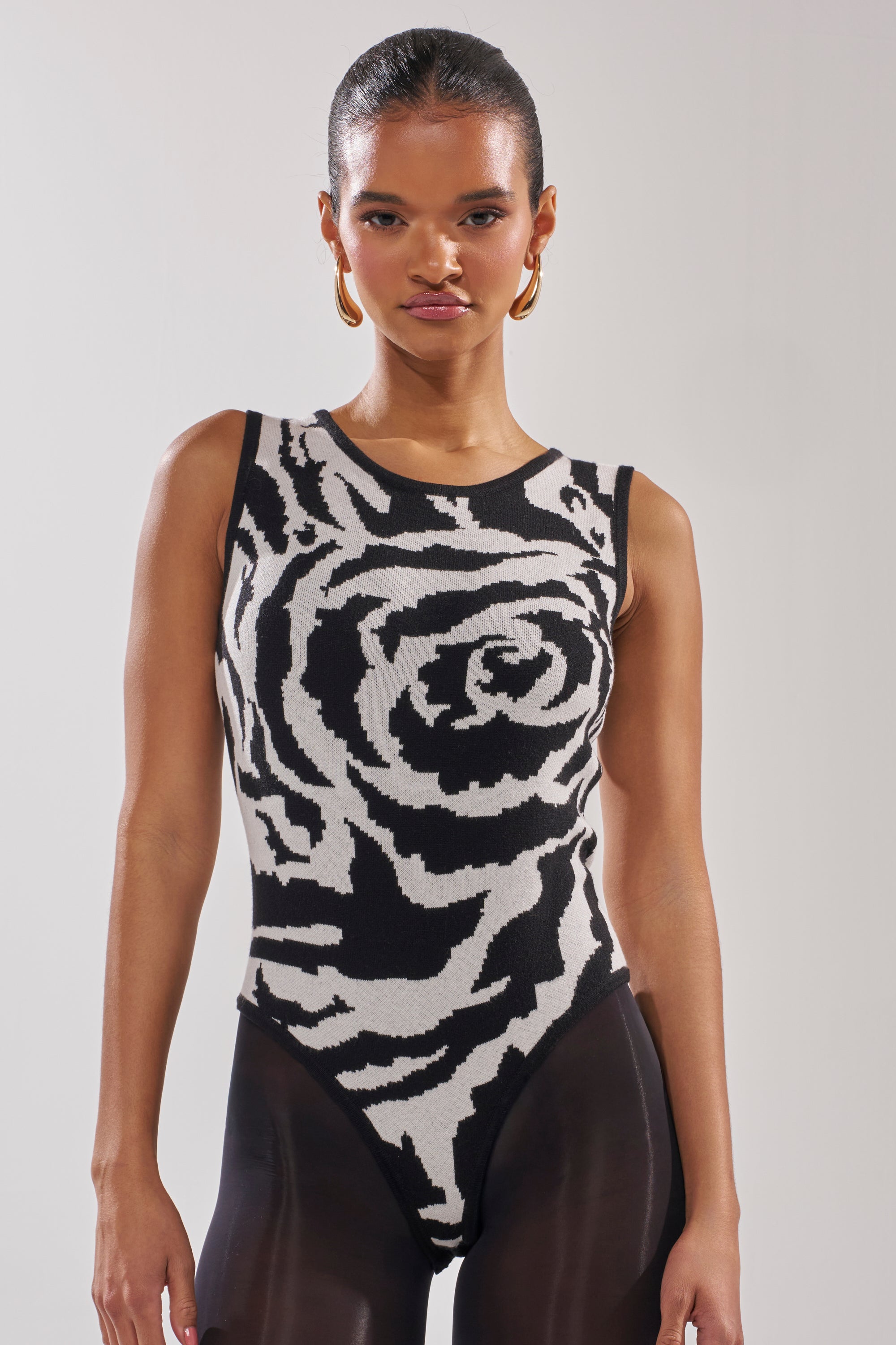 A woman in the NATOMA FLORAL BODYSUIT, featuring a bold black-and-white rose print and stretchy knit fabric, pairs it with sheer black leggings as she stands against a plain light background.