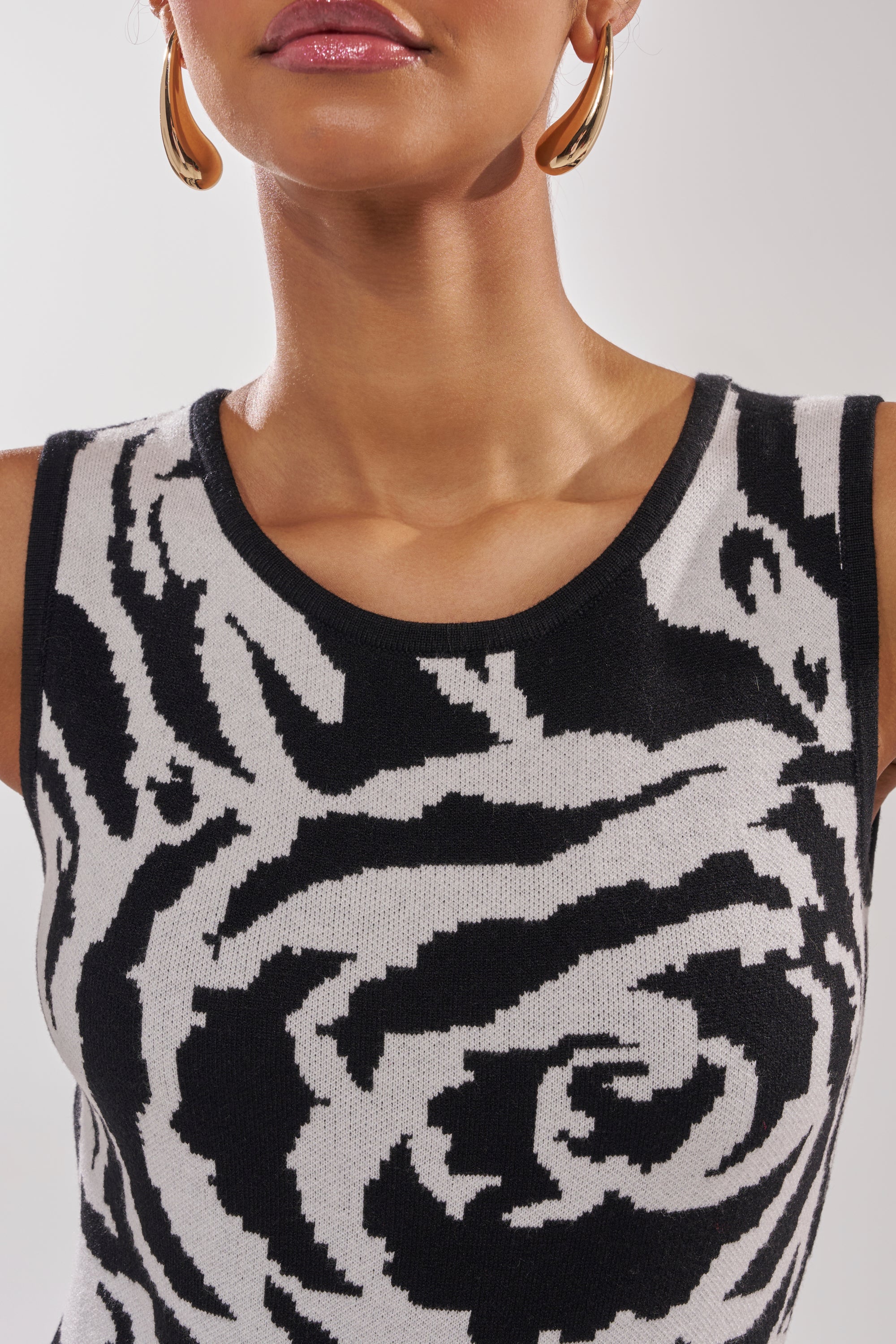 A woman wears gold hoop earrings and a sleeveless NATOMA FLORAL BODYSUIT with a bold black and white rose print. The image is cropped from lips to chest.