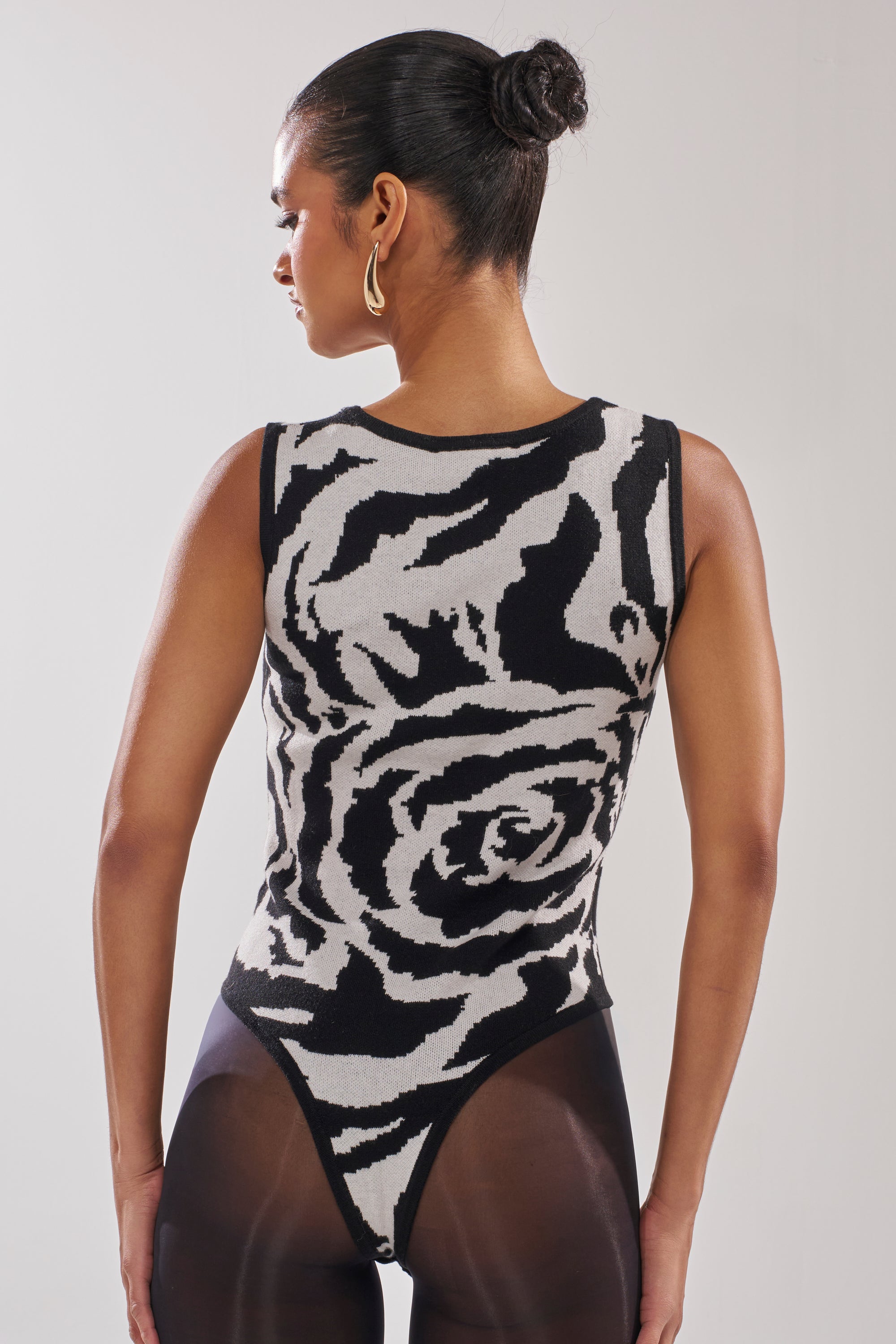 A woman with her hair in a bun and gold hoop earrings wears the NATOMA FLORAL BODYSUIT and sheer black tights, facing away to show the back of her outfit.