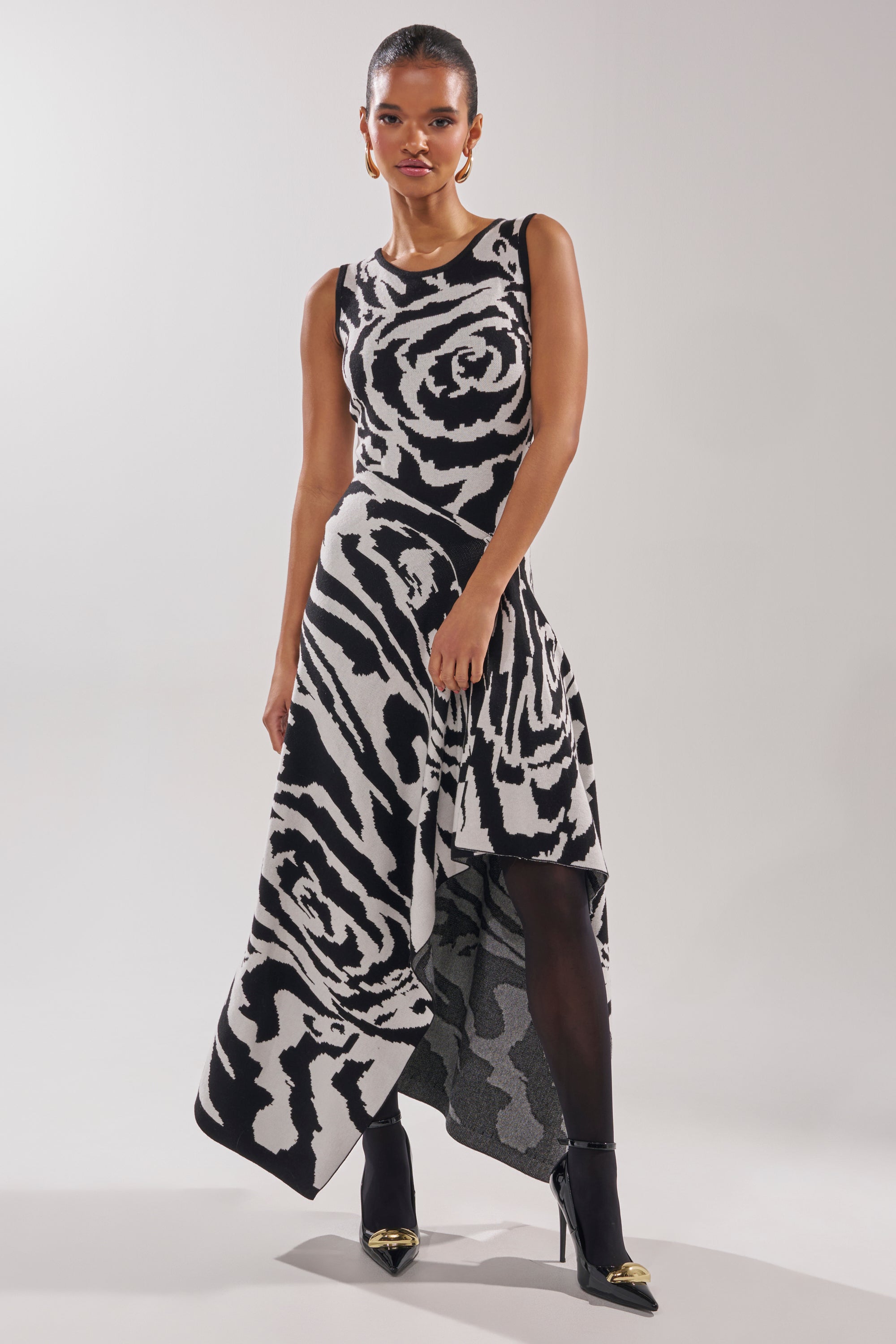 A woman wears the NATOMA FLORAL BODYSUIT with a striking black and white rose print, paired with black tights and heels, standing against a plain light background.