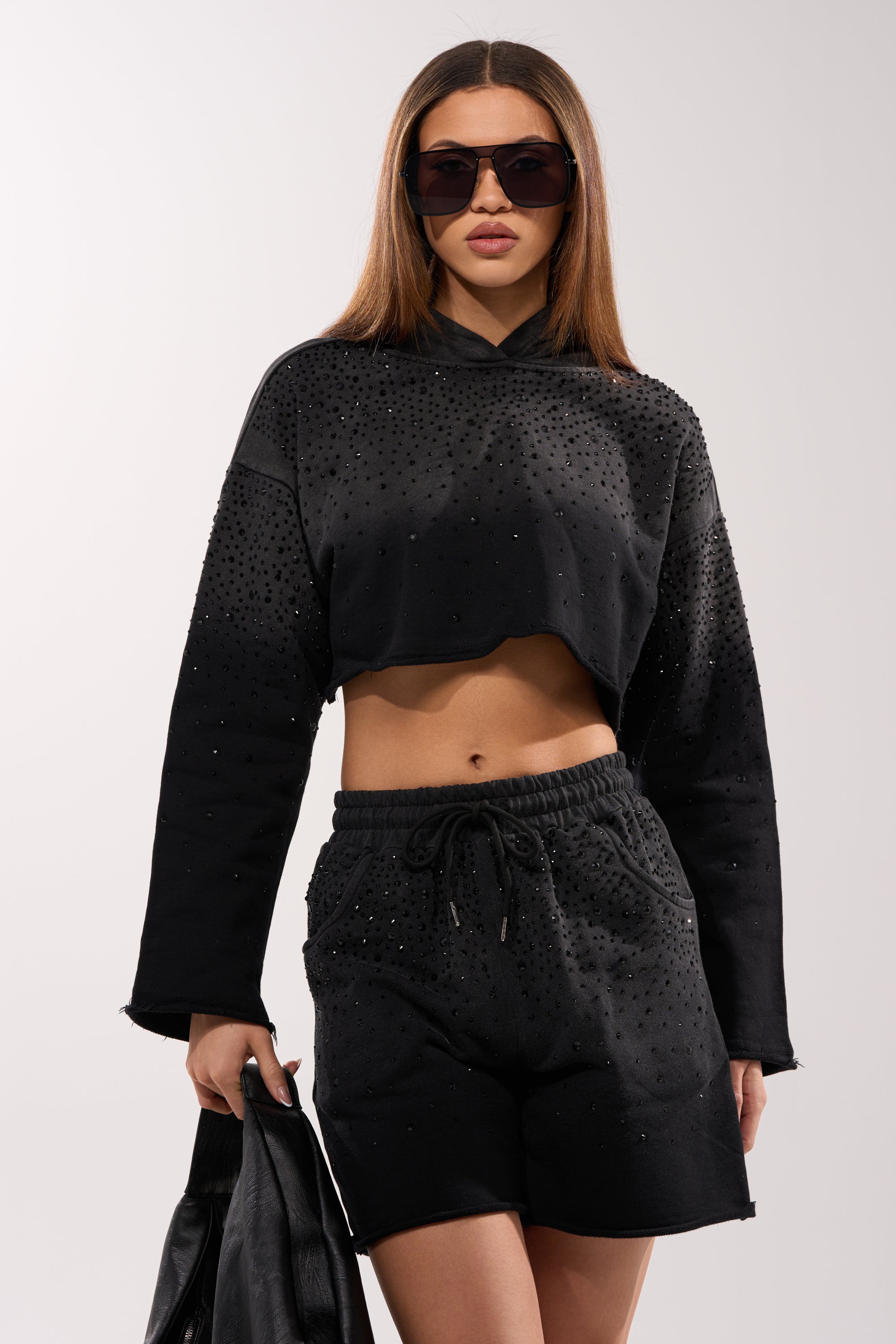 A woman wears the SPARKLE AND SHINE MINERAL WASHED CROPPED HOODIE with matching studded shorts, holding a black jacket. She has straight brown hair, large sunglasses, and sports a trendy casual look. 0