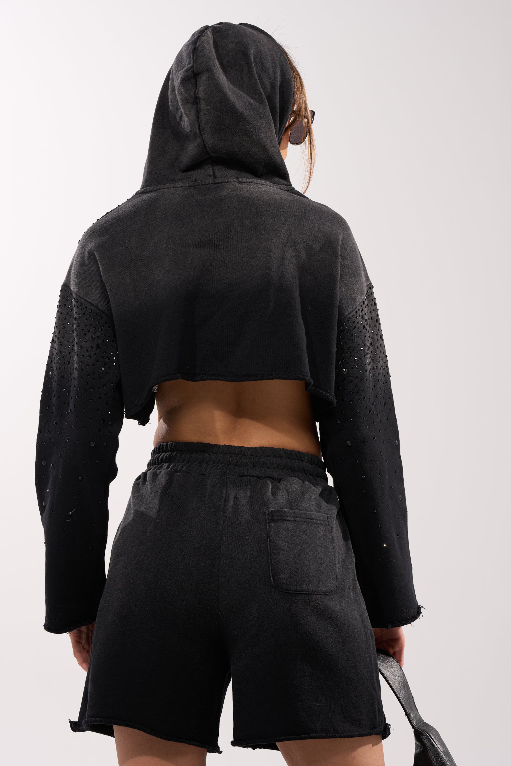 Someone wearing the SPARKLE AND SHINE MINERAL WASHED CROPPED HOODIE with the hood up and matching black shorts, featuring embellished sleeves and a sporty design; the shorts include a back pocket. 4