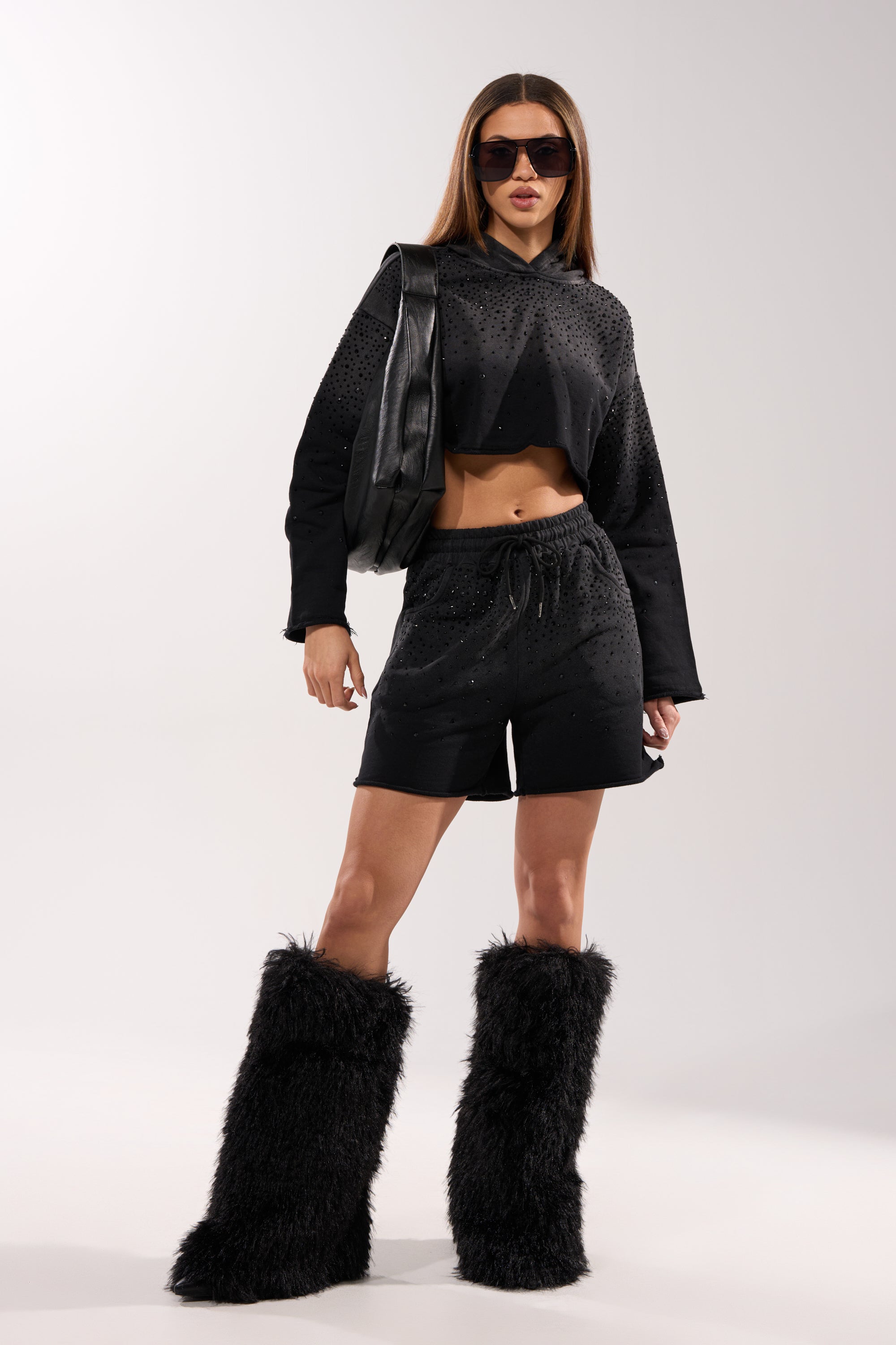 A woman stands confidently in the SPARKLE AND SHINE MINERAL WASHED CROPPED HOODIE, black shorts, oversized sunglasses, a black shoulder bag, and tall furry black boots against a plain light background. 5