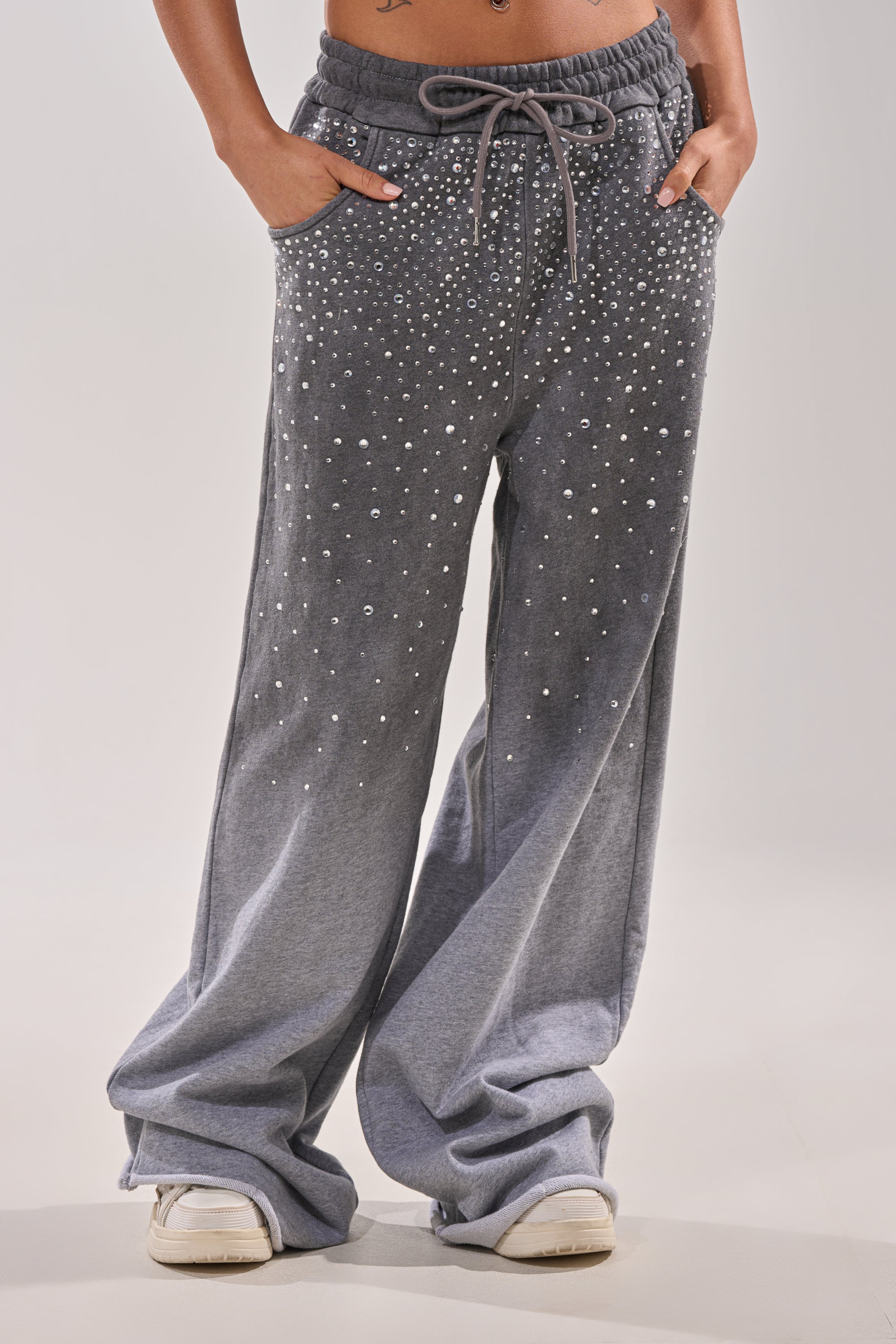 A person wears the NIGHT SKY RHINESTONE JOGGER, featuring a wide-leg grey design with scattered pearl-like rhinestone embellishments. Hands are in pockets, paired with white sneakers. The image is cropped from the waist down. 1