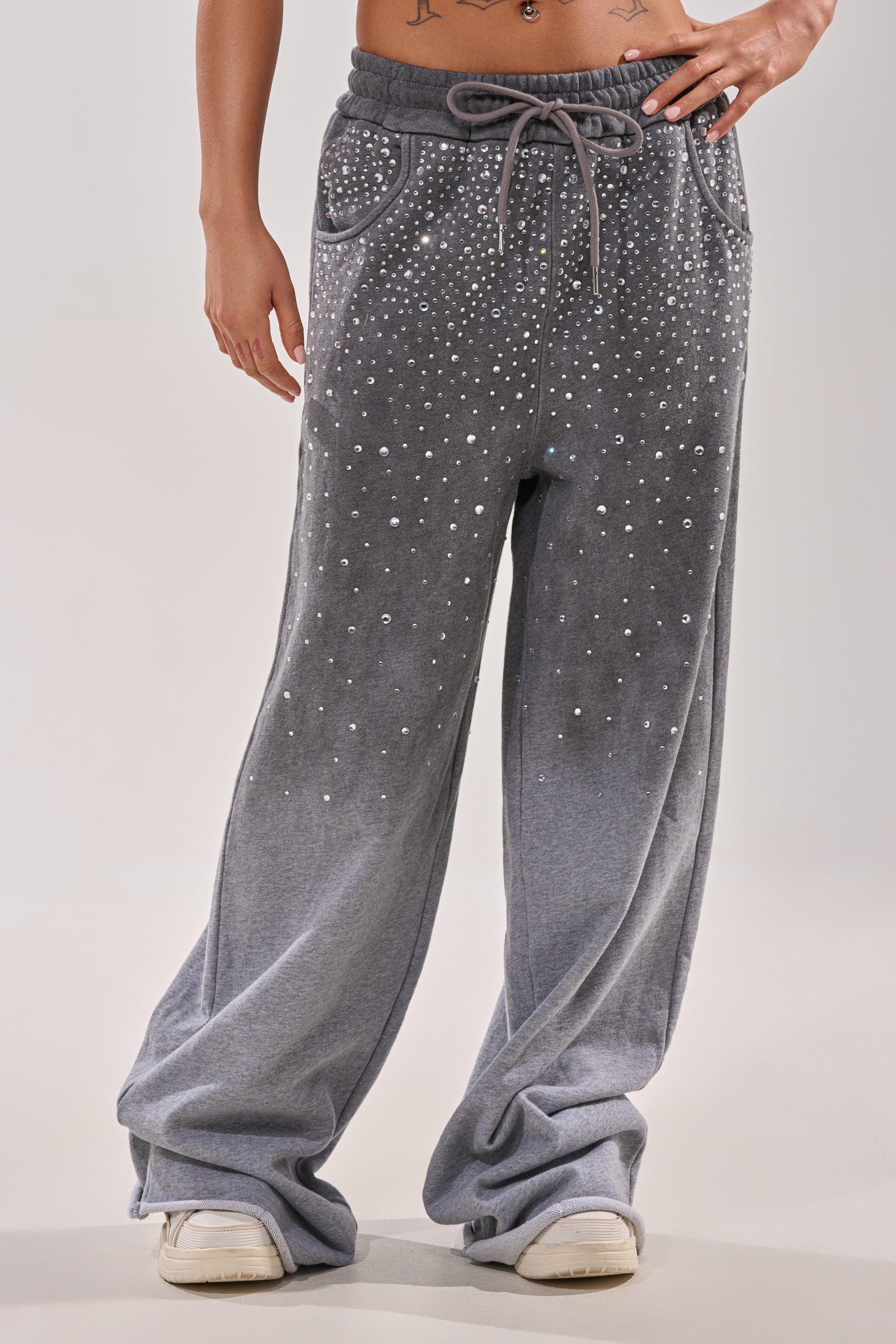 A person models the NIGHT SKY RHINESTONE JOGGER—loose-fitting grey joggers with pearl-like rhinestone details—paired with white sneakers, standing against a plain, light backdrop. 2