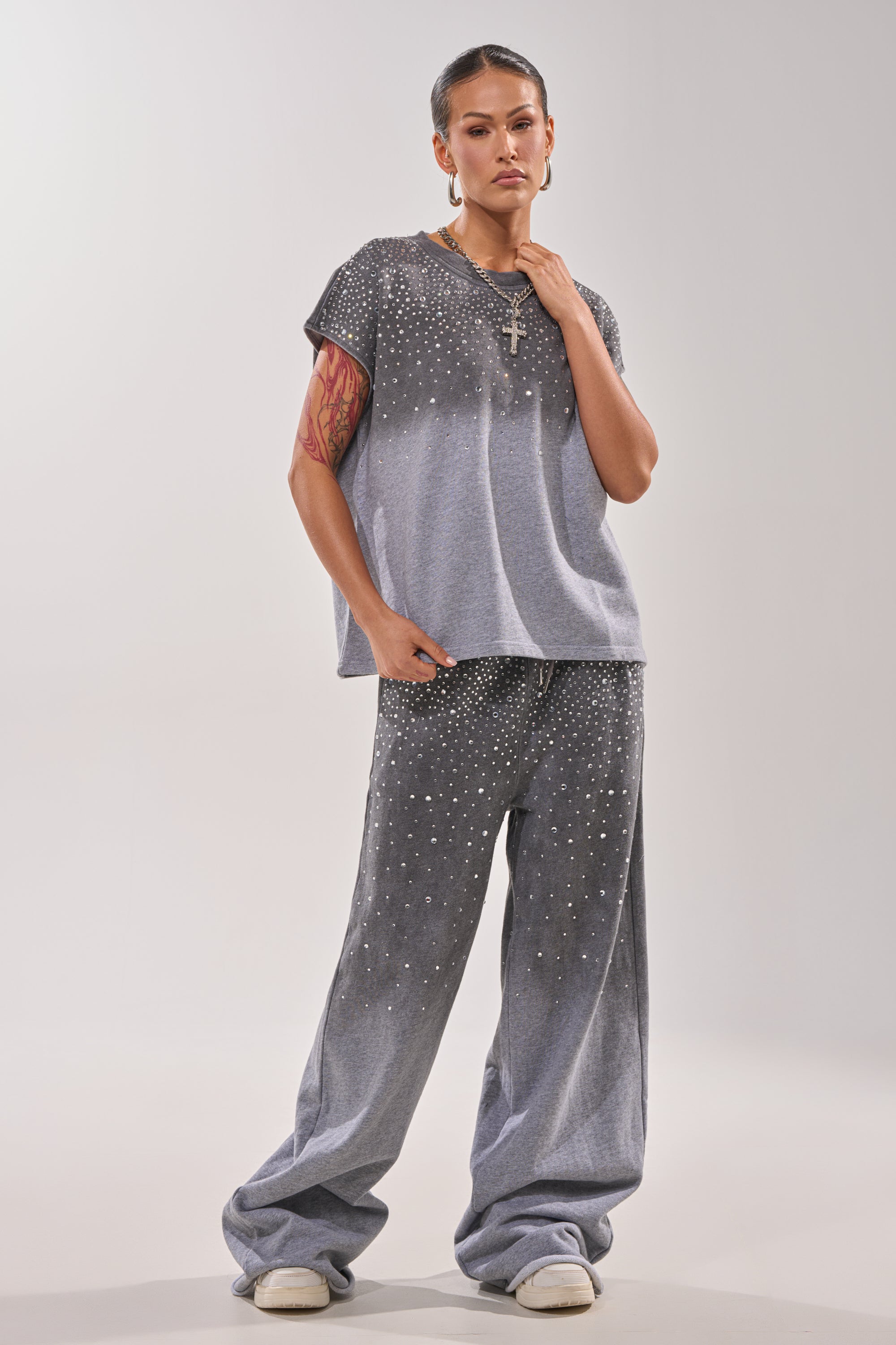 A person stands against a plain background wearing the NIGHT SKY RHINESTONE JOGGER, a loose, grey, short-sleeve top and matching wide-leg joggers adorned with small white dots. They accessorize with tattoos, jewelry, and white sneakers. 5
