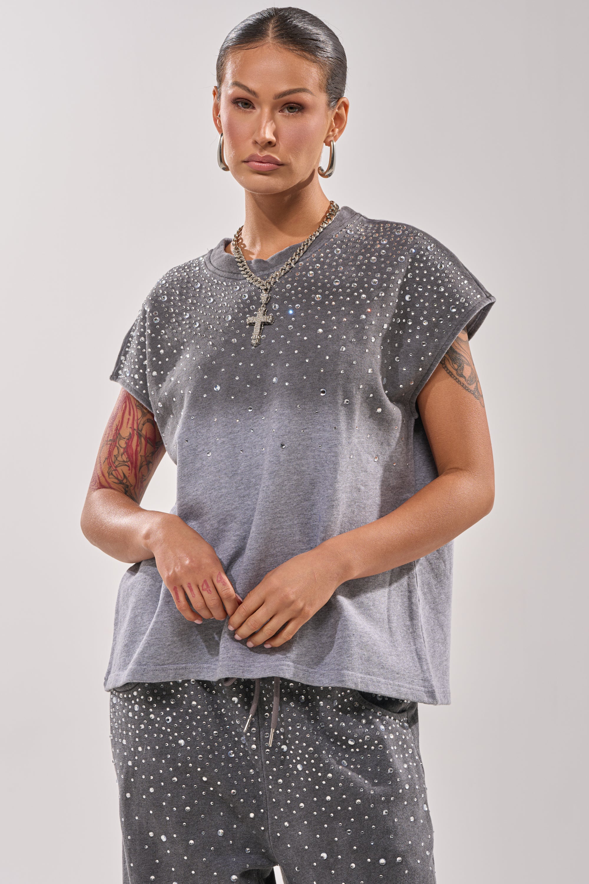 A person with tattoos wears the casual NIGHT SKY RHINESTONE TEE, featuring a lightweight gray knit and small silver studs, paired with matching pants, gold hoop earrings and necklace, and slicked-back hair. 0